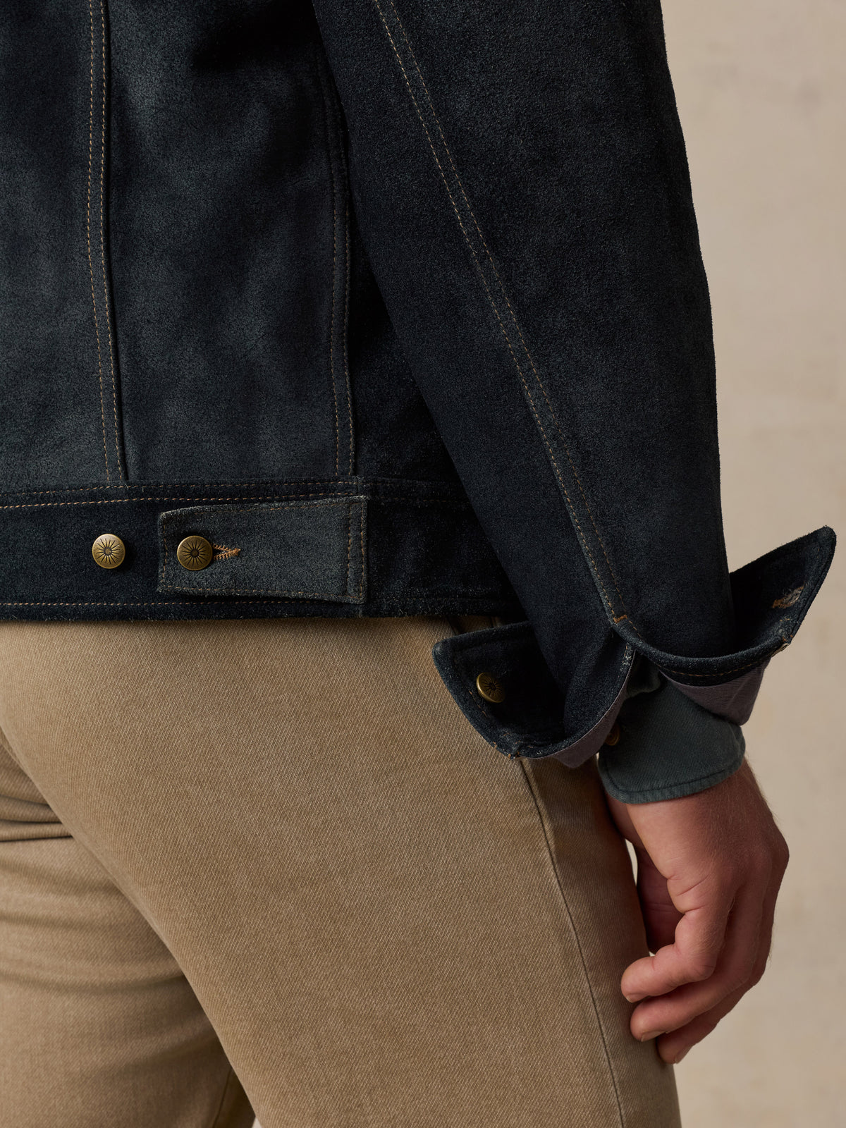 Close-up of a man wearing a dark indigo denim jacket with rolled cuffs and tan pants, showing the sleeve, button details, and hand against a neutral beige background.