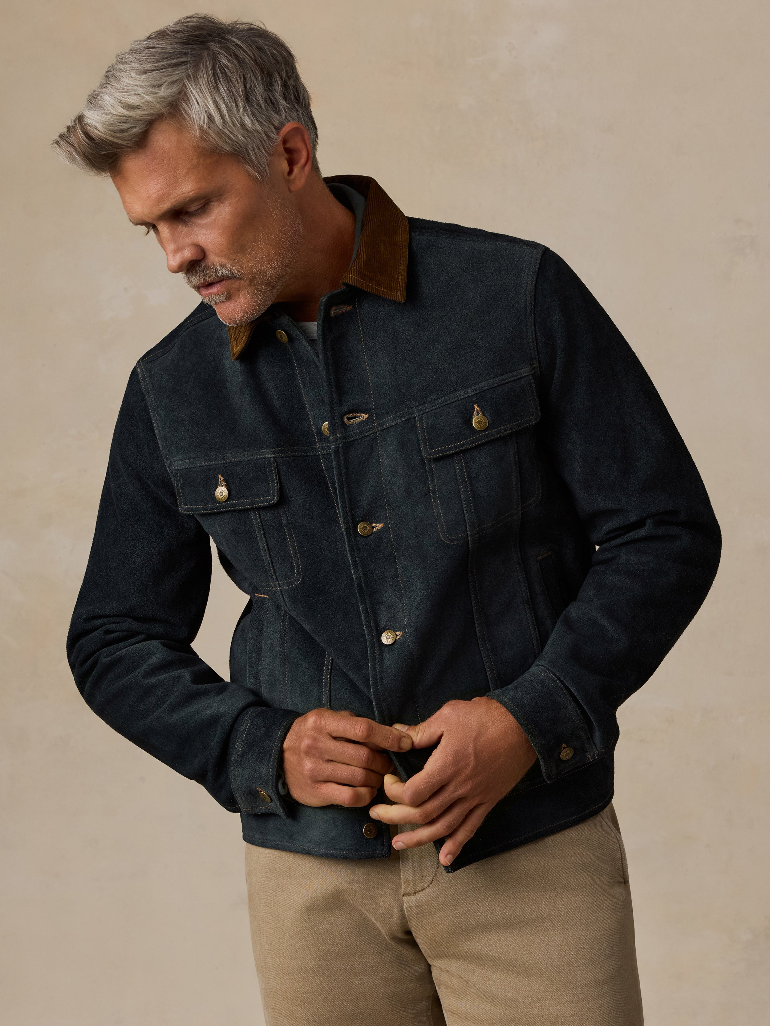 A man wearing a dark indigo denim jacket with a brown corduroy collar and brass buttons, paired with beige trousers, standing against a textured beige background.