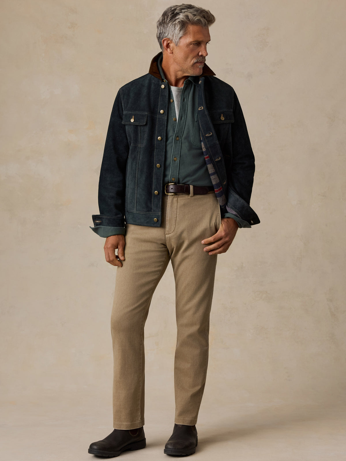 A man stands against a textured beige studio backdrop wearing a dark indigo denim jacket with a brown corduroy collar, a green shirt, khaki pants, and dark brown boots.