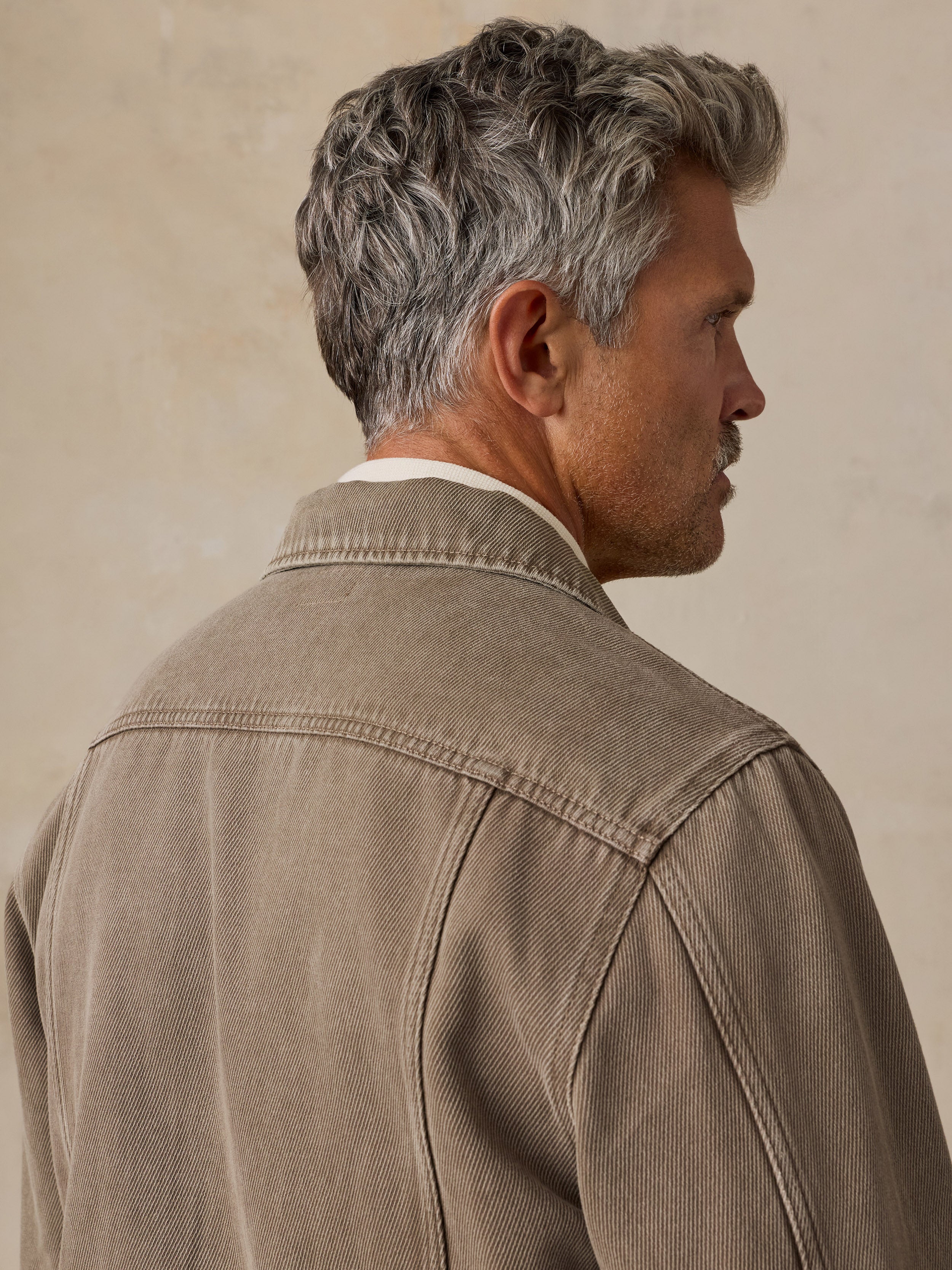 A man shown from the back and side profile wearing a textured taupe jacket over a white collared shirt, set against a warm beige background.