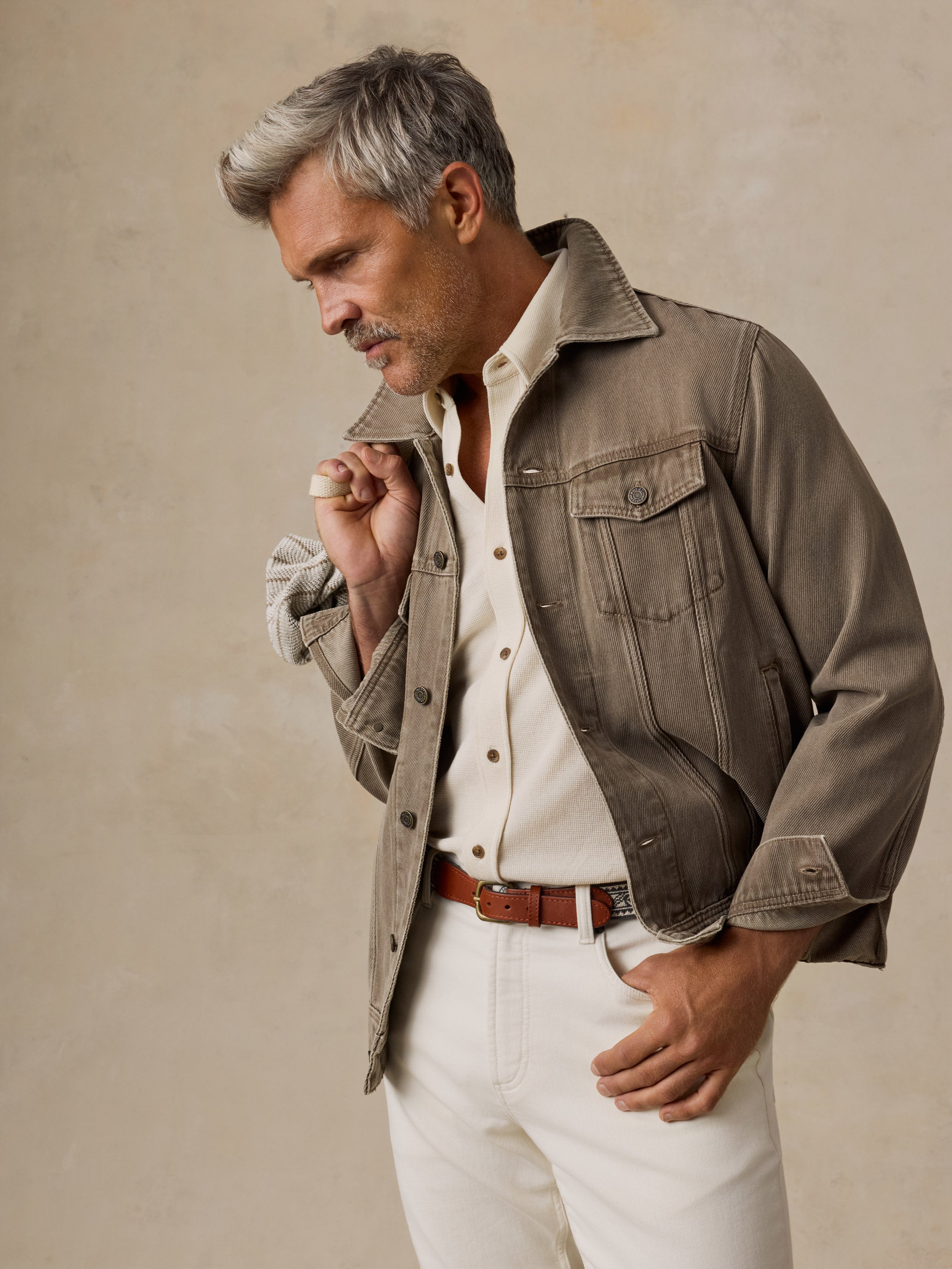 A man stands against a textured beige studio backdrop wearing a taupe jacket over a cream shirt and white trousers