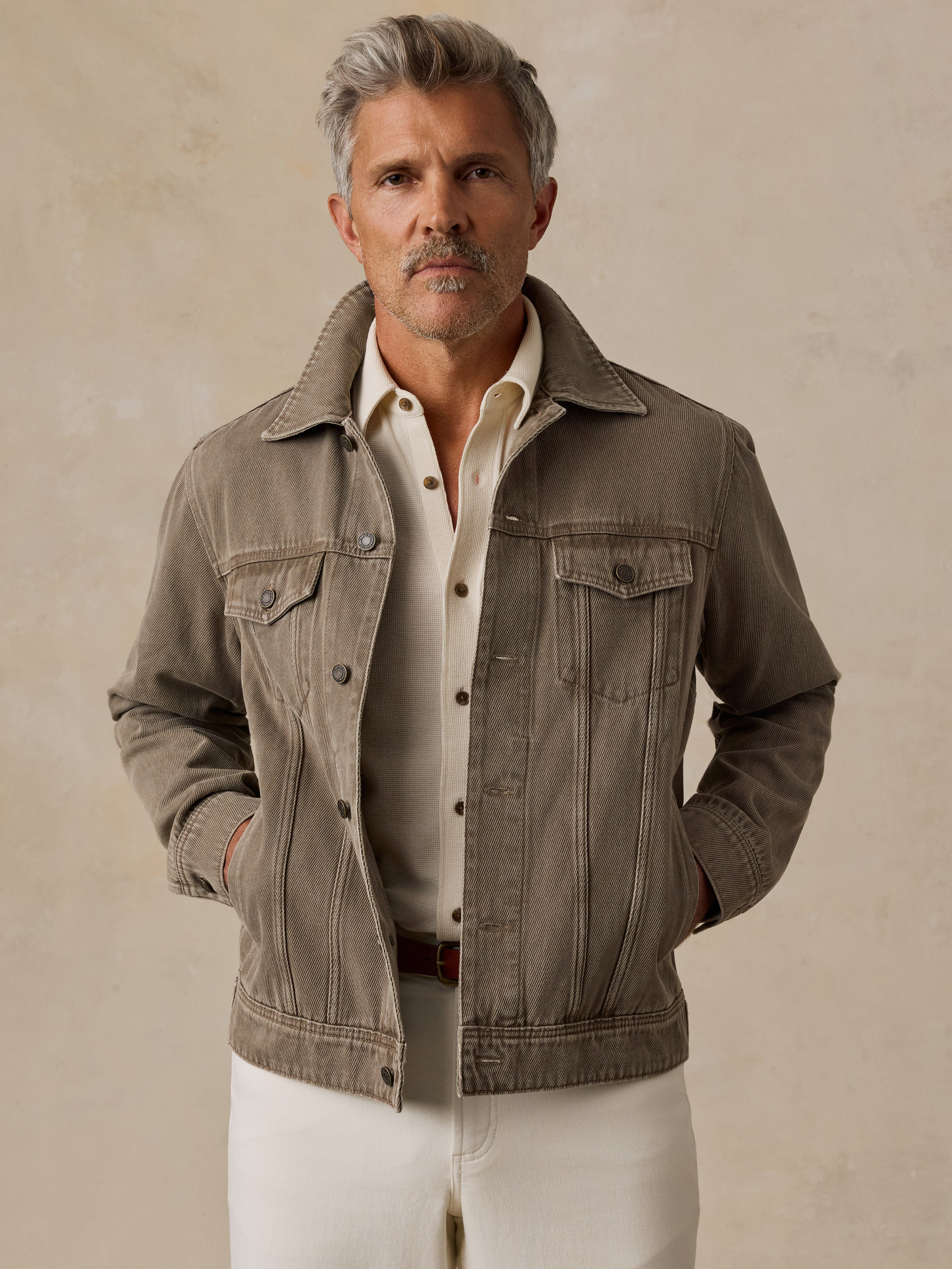 A man stands against a textured beige studio backdrop wearing a taupe jacket over a cream shirt and white trousers