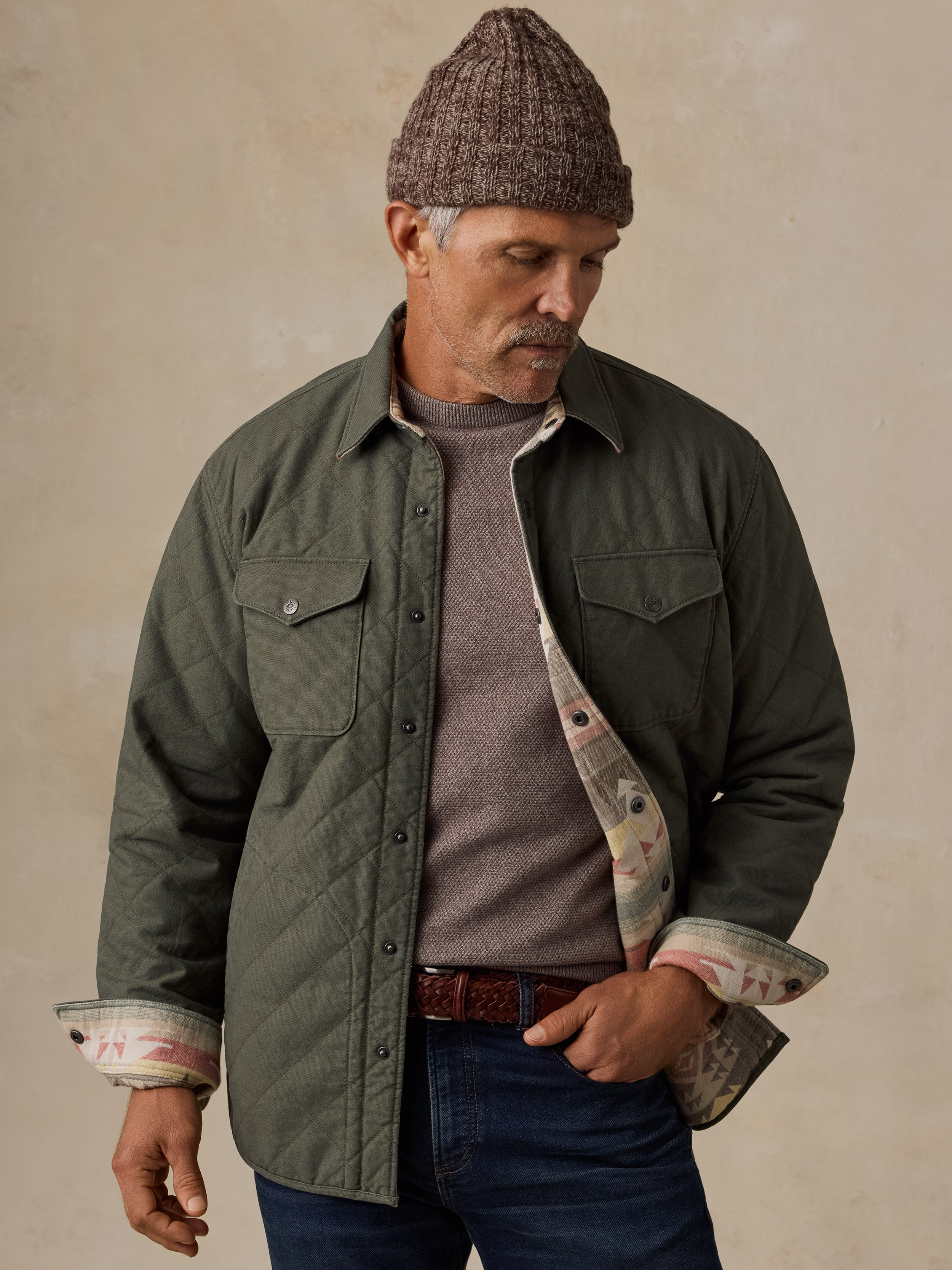 A man wearing an olive green quilted button-up jacket with a patterned lining, a brown knit beanie, a gray t-shirt, and blue jeans, standing against a textured beige background.