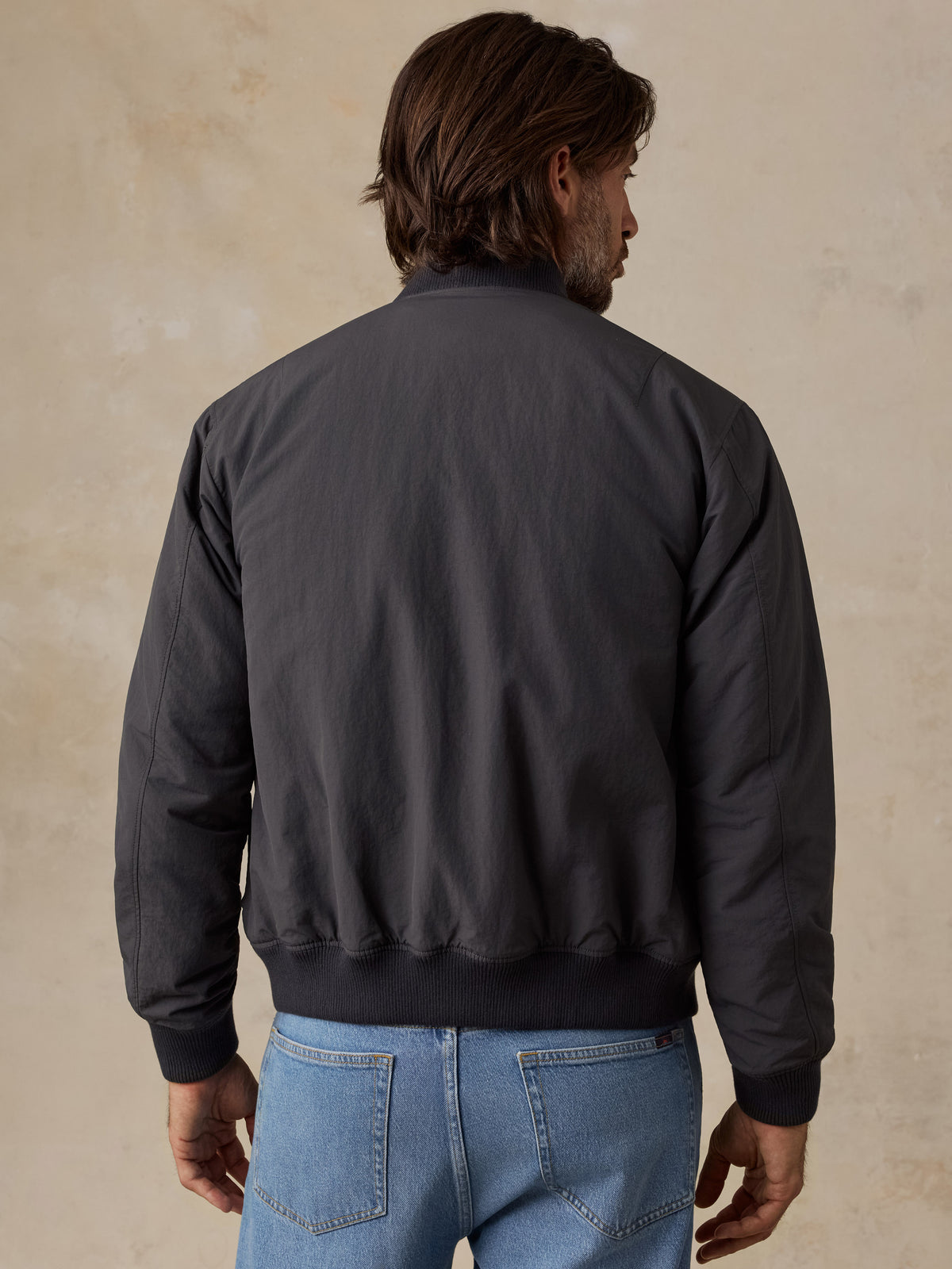 A man viewed from behind wearing a dark charcoal gray bomber jacket and light blue jeans, standing against a textured beige background., image 4 of 6