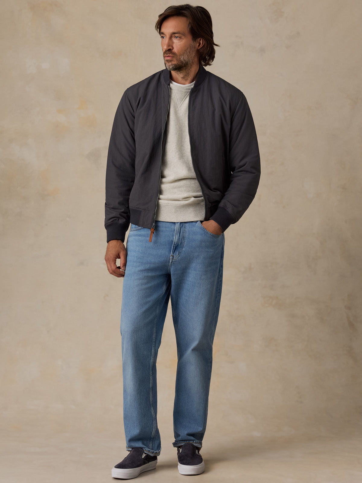 A man wearing a dark gray bomber jacket, a light gray crewneck sweater, blue jeans, and dark sneakers, standing against a textured beige studio backdrop., image 2 of 6