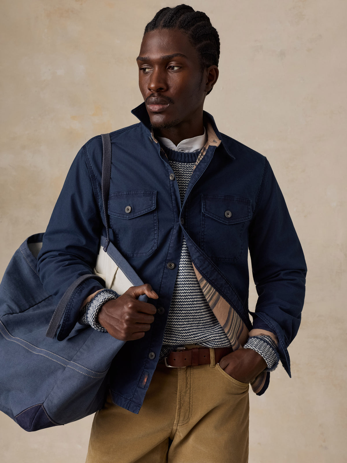 A man wearing a blue jacket over a blue striped sweater and tan corduroy pants holding a blue tote bag, image 5 of 5