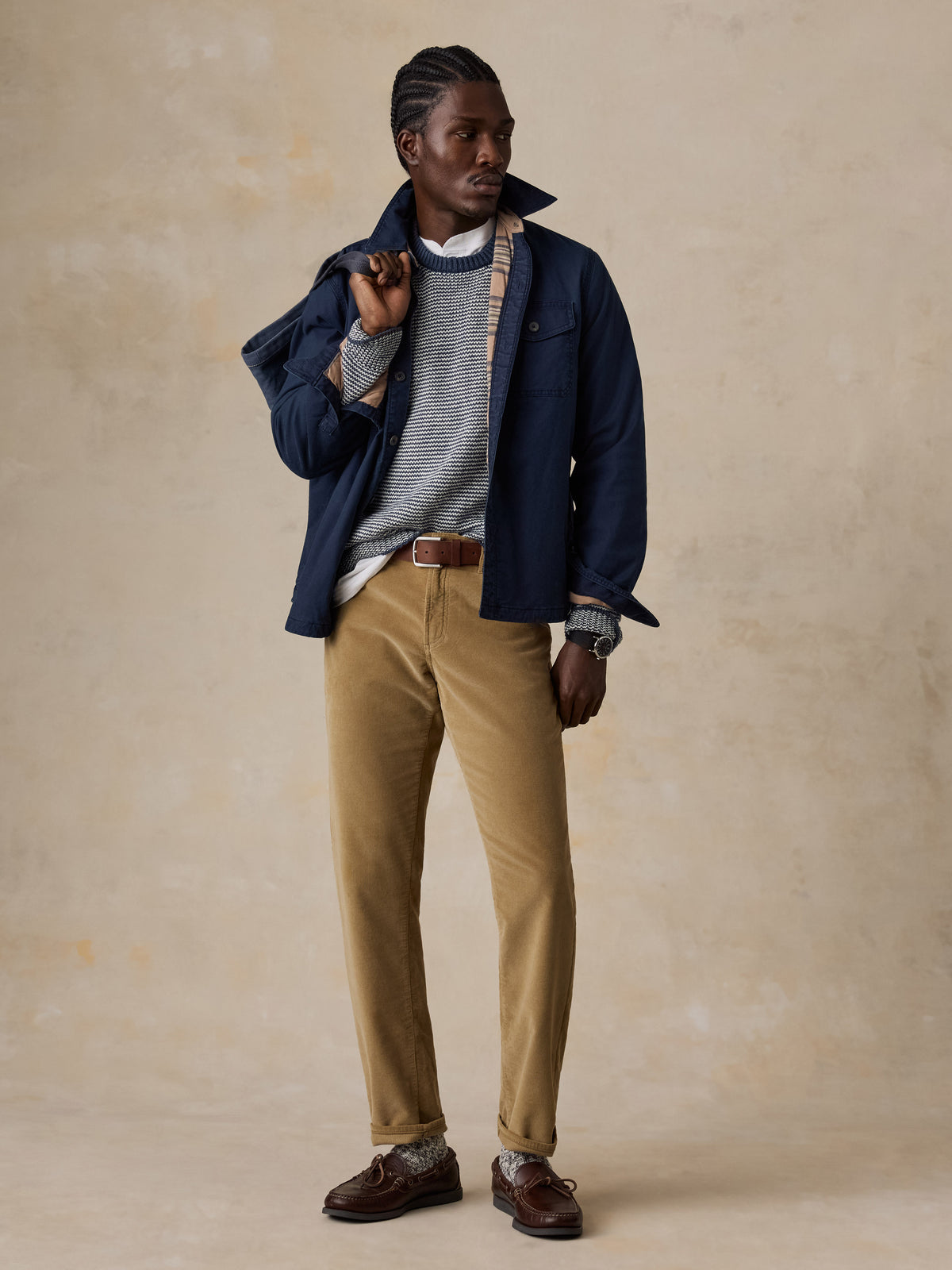 A man wearing a blue jacket over a blue striped sweater and tan corduroy pants holding a blue tote bag, image 2 of 5