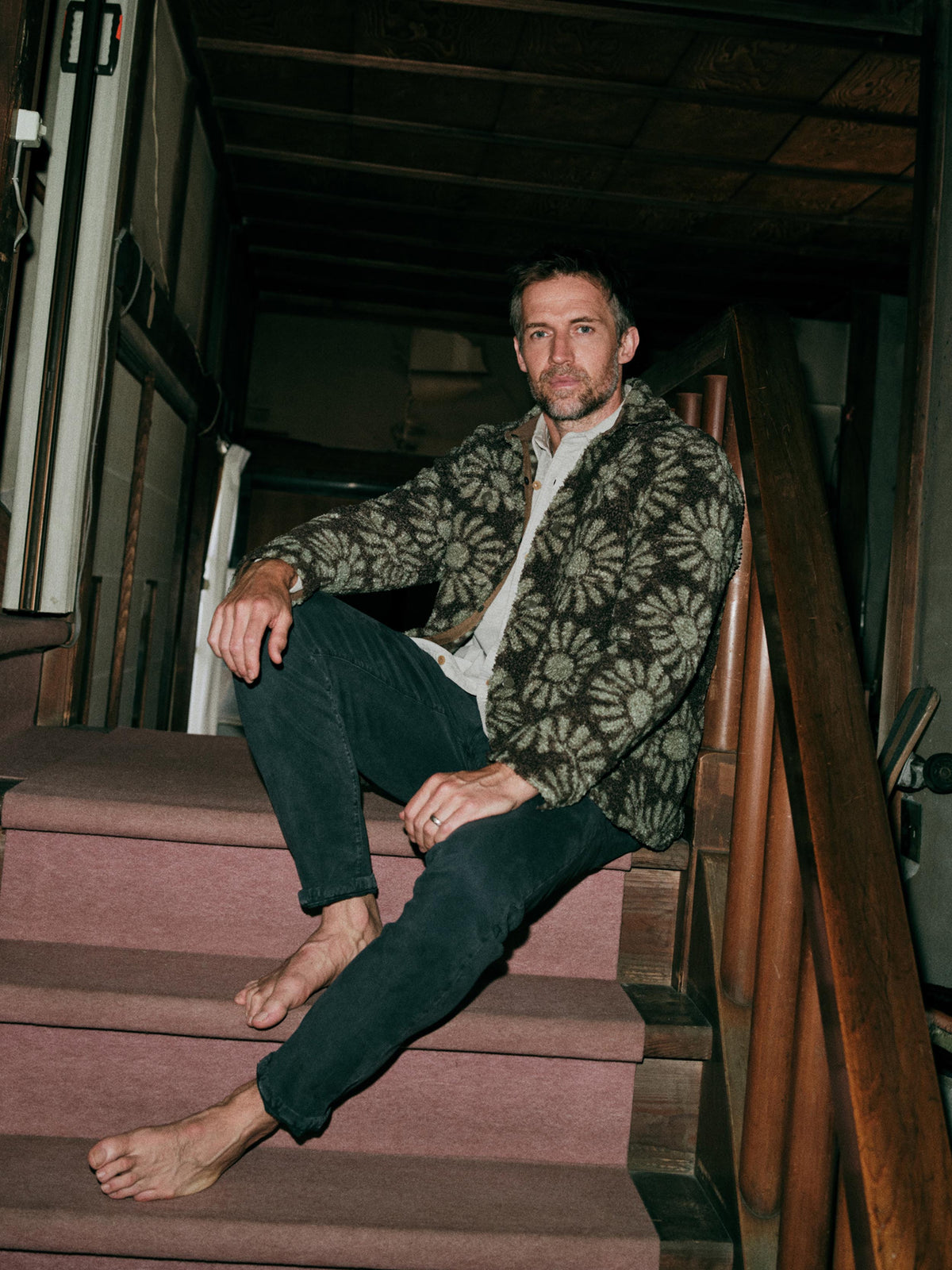 A man sits on a wooden staircase indoors, wearing a floral patterned fleece jacket over a button up shirt and dark pants., image 1 of 7