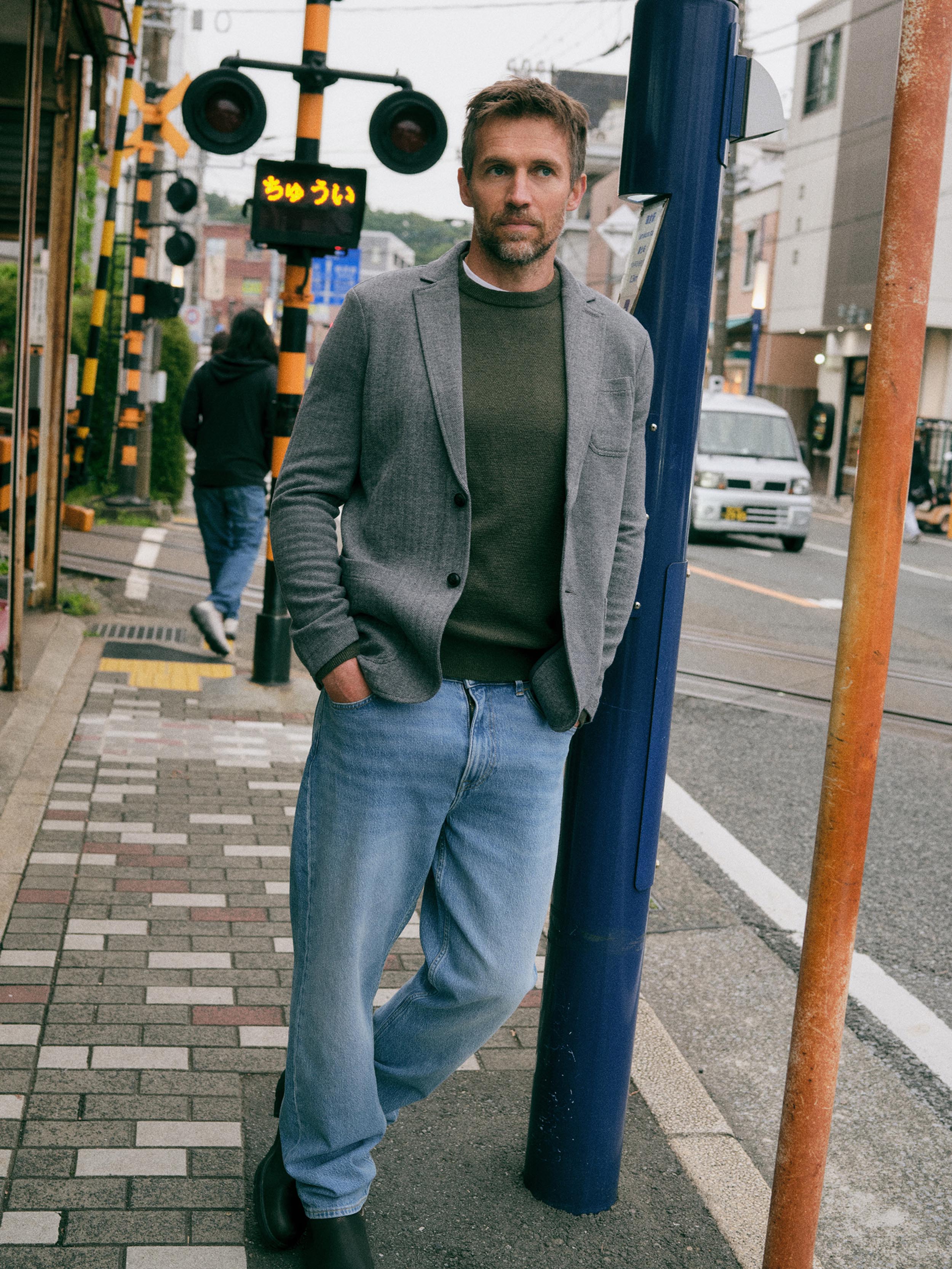 A man stands on a city sidewalk wearing a gray blazer over a dark olive green sweater, light blue jeans, and black boots, leaning against a blue pole in an urban street setting.