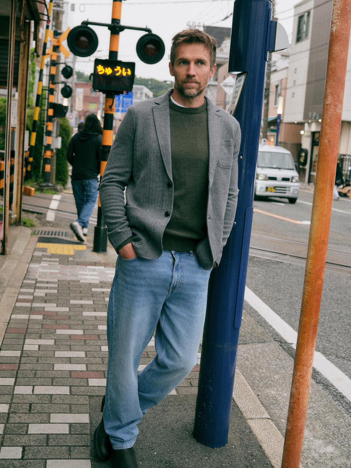 A man stands on a city sidewalk wearing a gray blazer over a dark olive green sweater, light blue jeans, and black boots, leaning against a blue pole in an urban street setting., image 1 of 7