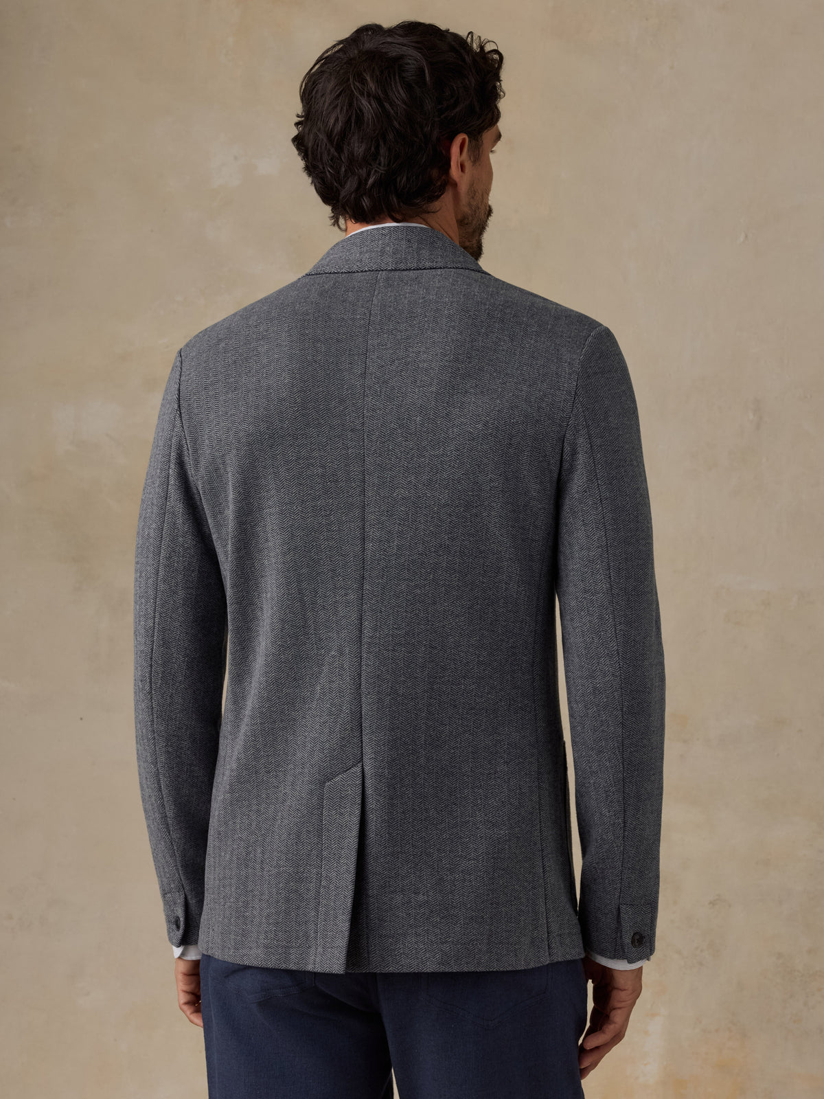 A man viewed from the back wearing a dark gray textured blazer and navy trousers, standing against a warm beige textured wall., image 5 of 7