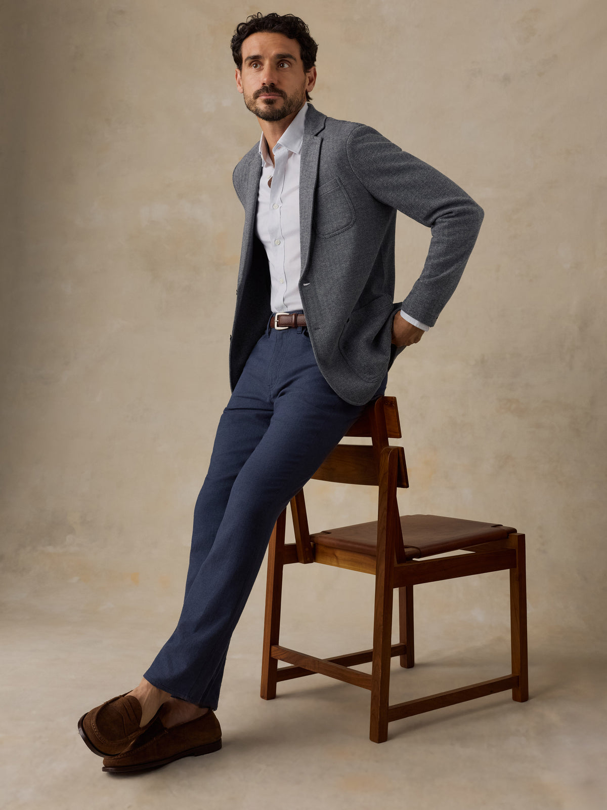 A man wearing a gray blazer, white shirt, navy trousers, and brown loafers leans against a wooden chair in a studio setting with a textured beige background., image 6 of 7