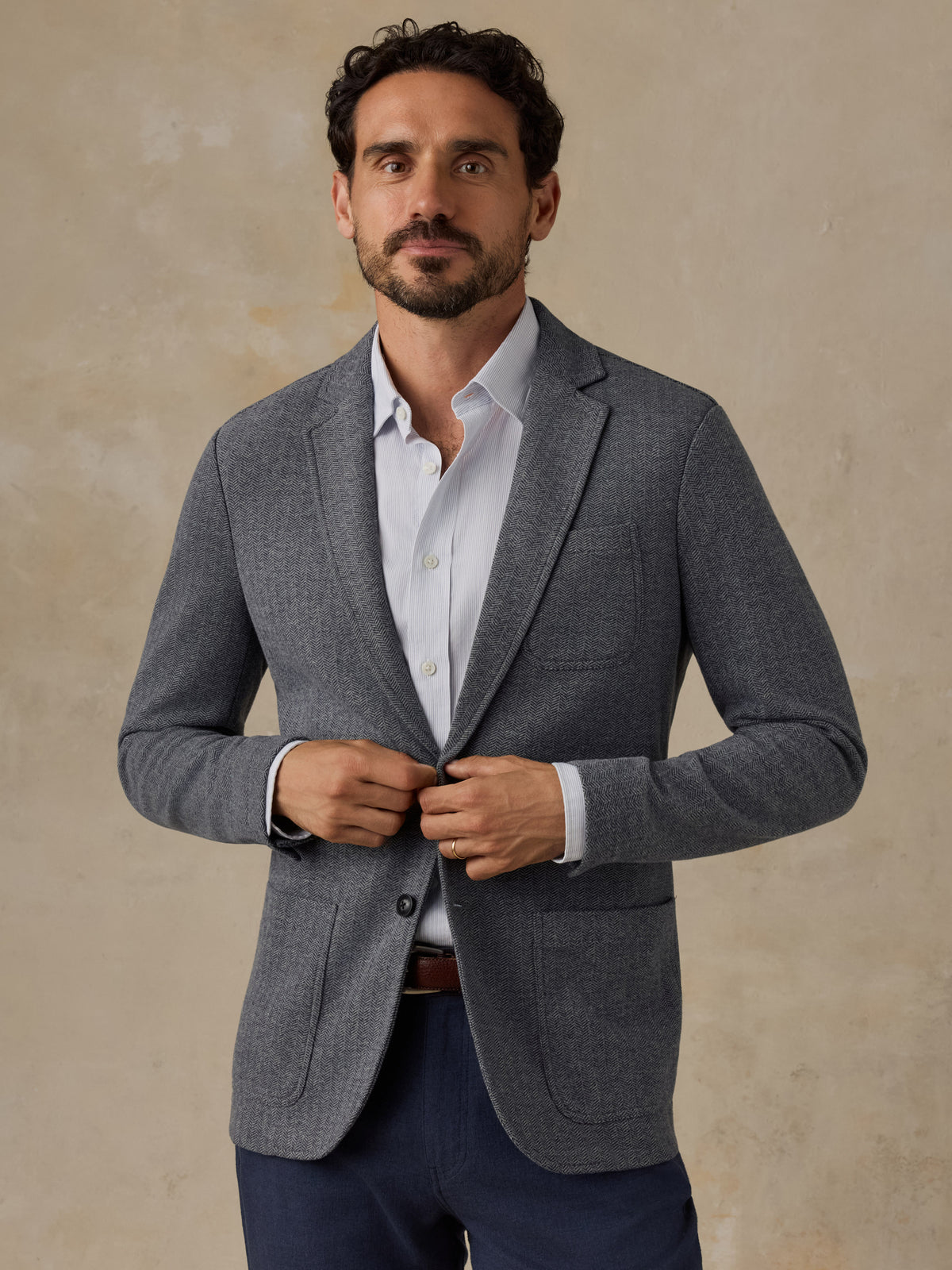 A man wearing a gray textured blazer over a white dress shirt and navy trousers, standing against a textured beige background., image 2 of 7
