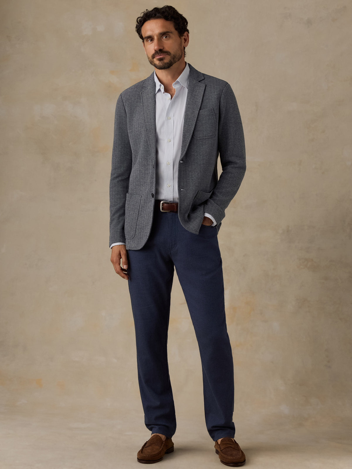 A man wearing a gray textured blazer, white dress shirt, navy trousers, and brown suede loafers, standing against a textured beige studio backdrop., image 4 of 7