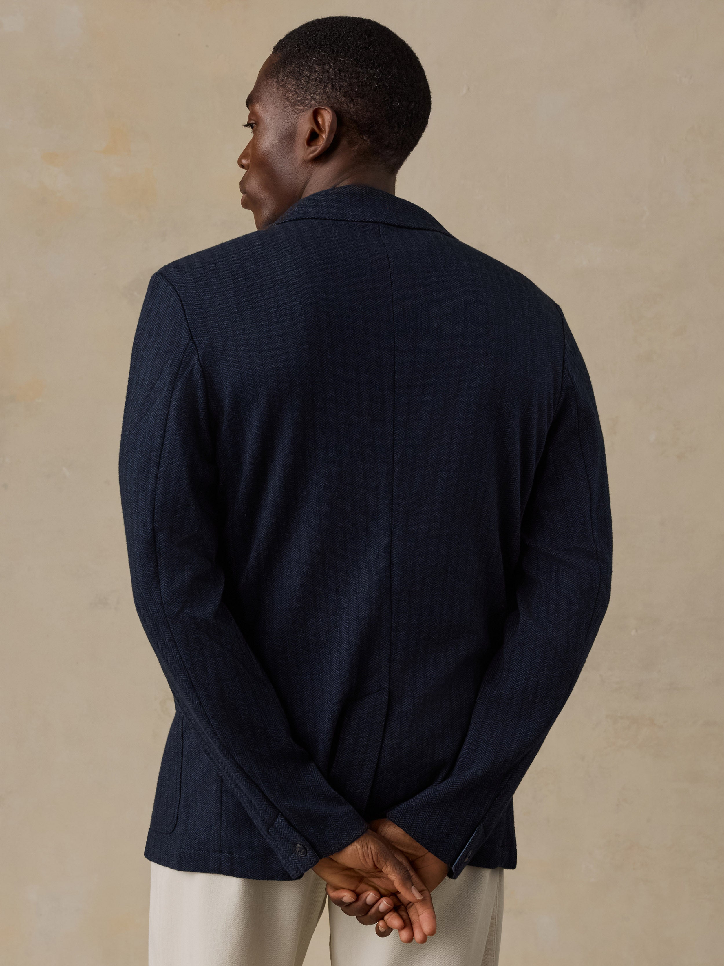 A man viewed from behind wearing a dark navy blue textured blazer and light beige trousers, standing against a textured beige wall.