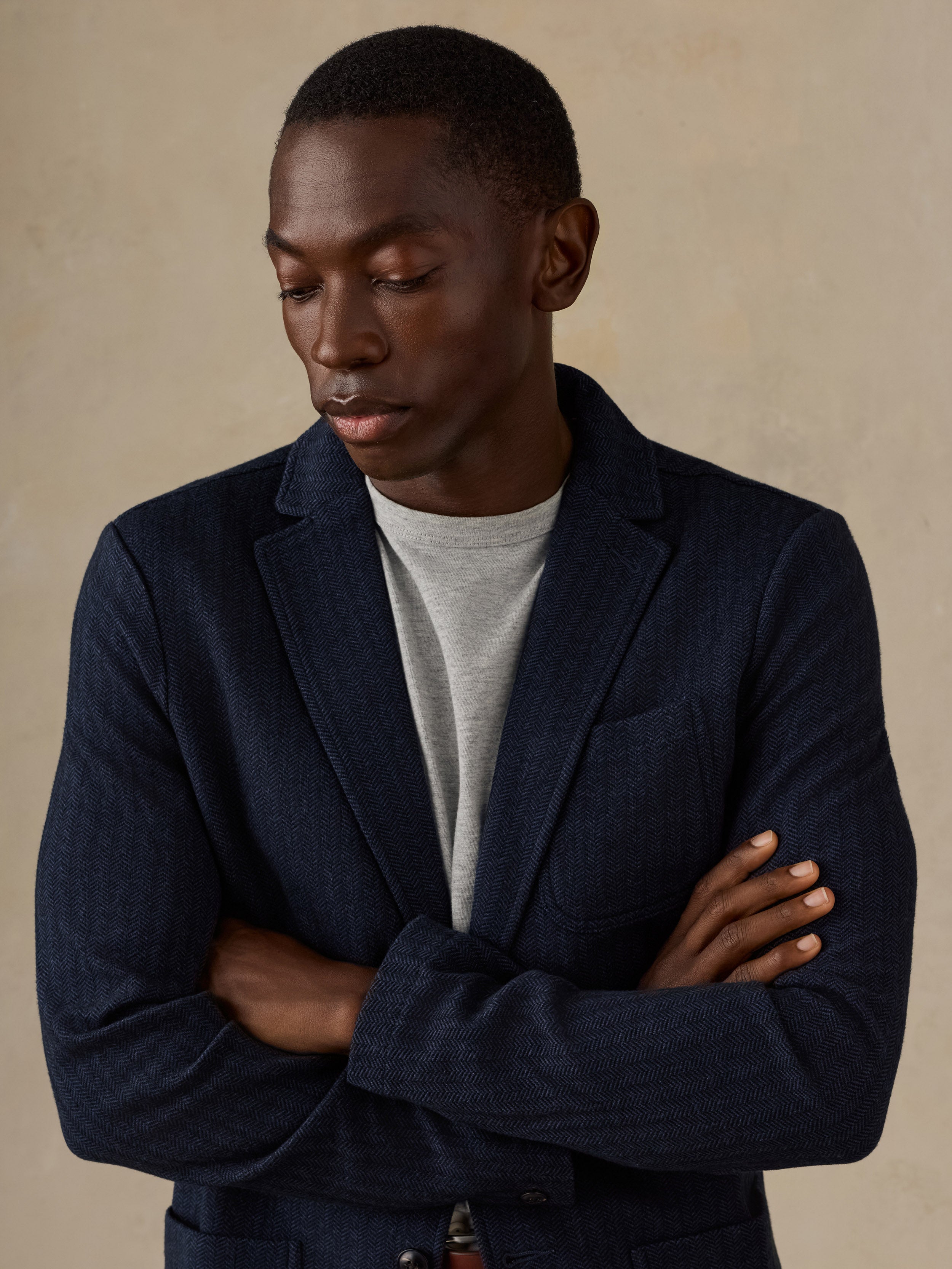A man wearing a dark navy textured blazer over a light gray crewneck t-shirt, standing with arms crossed against a warm beige background.