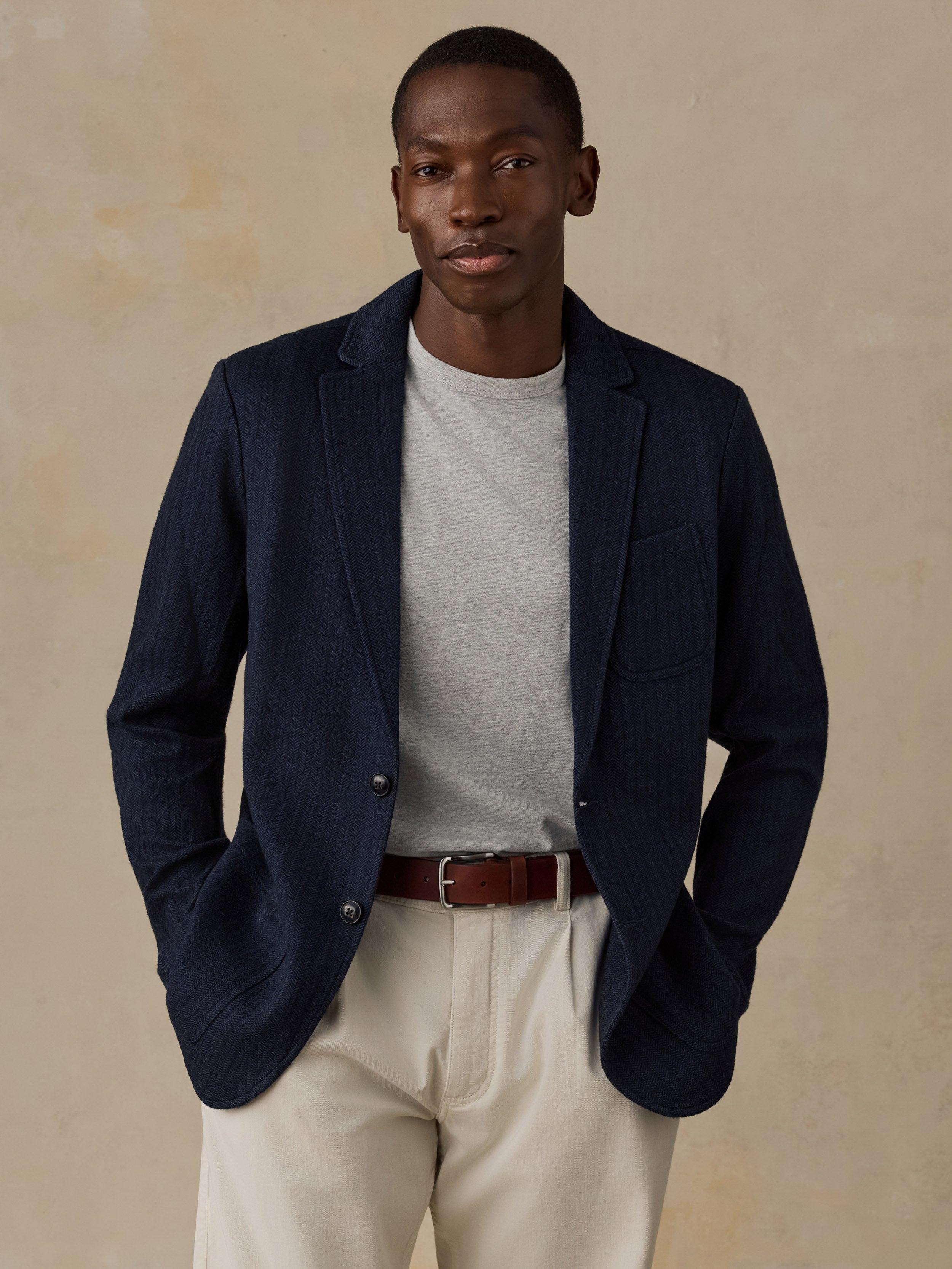 A man wearing a navy blue textured blazer over a light gray t-shirt, cream trousers, and a brown belt, standing against a textured beige background.