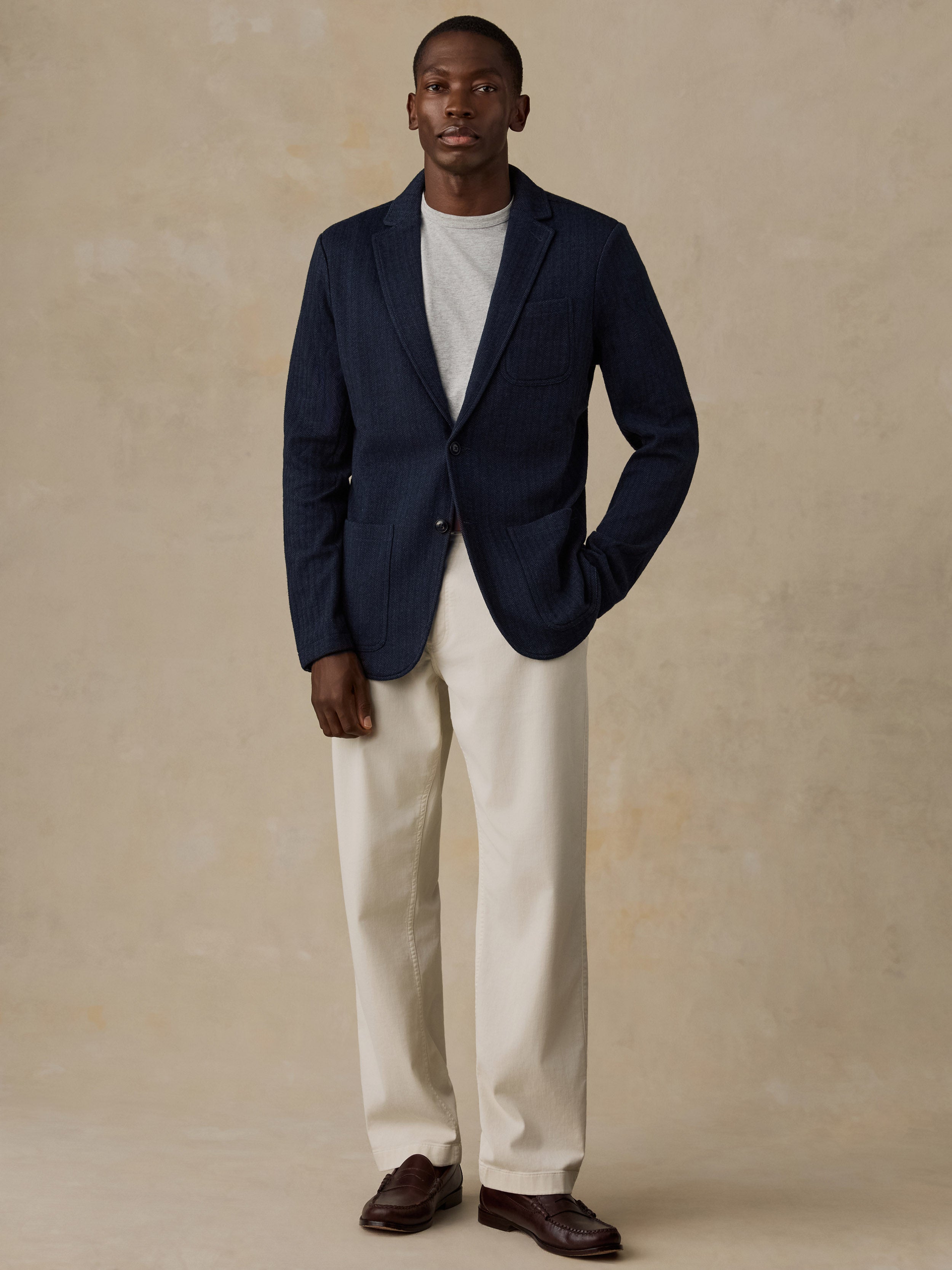A man stands against a textured beige studio backdrop wearing a navy blue pinstriped blazer over a light gray crewneck t-shirt, cream-colored trousers, and dark brown leather loafers.