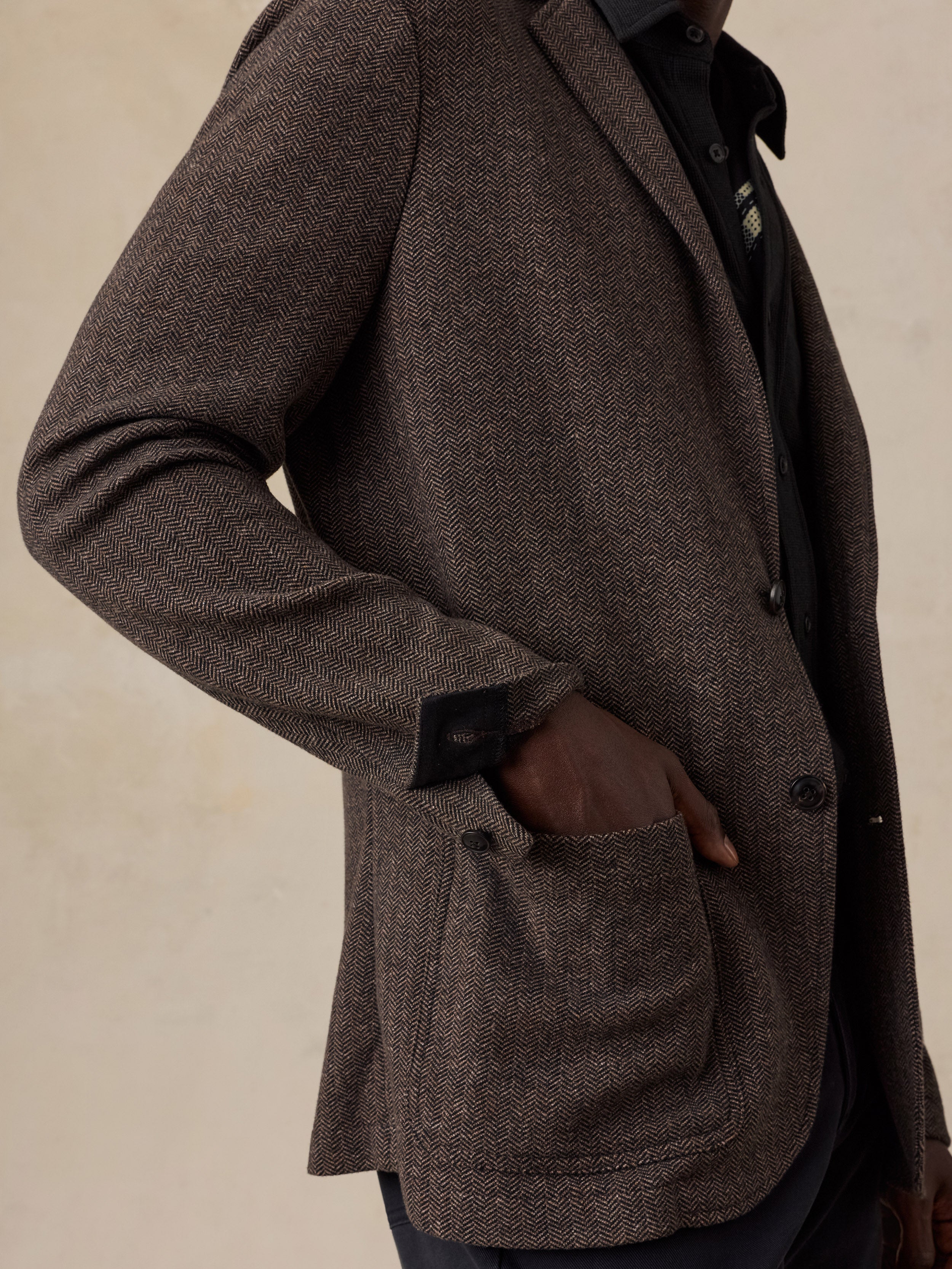 A close-up of a person man a textured brown herringbone blazer over a black collared shirt, with one hand in a front pocket, against a warm beige background.