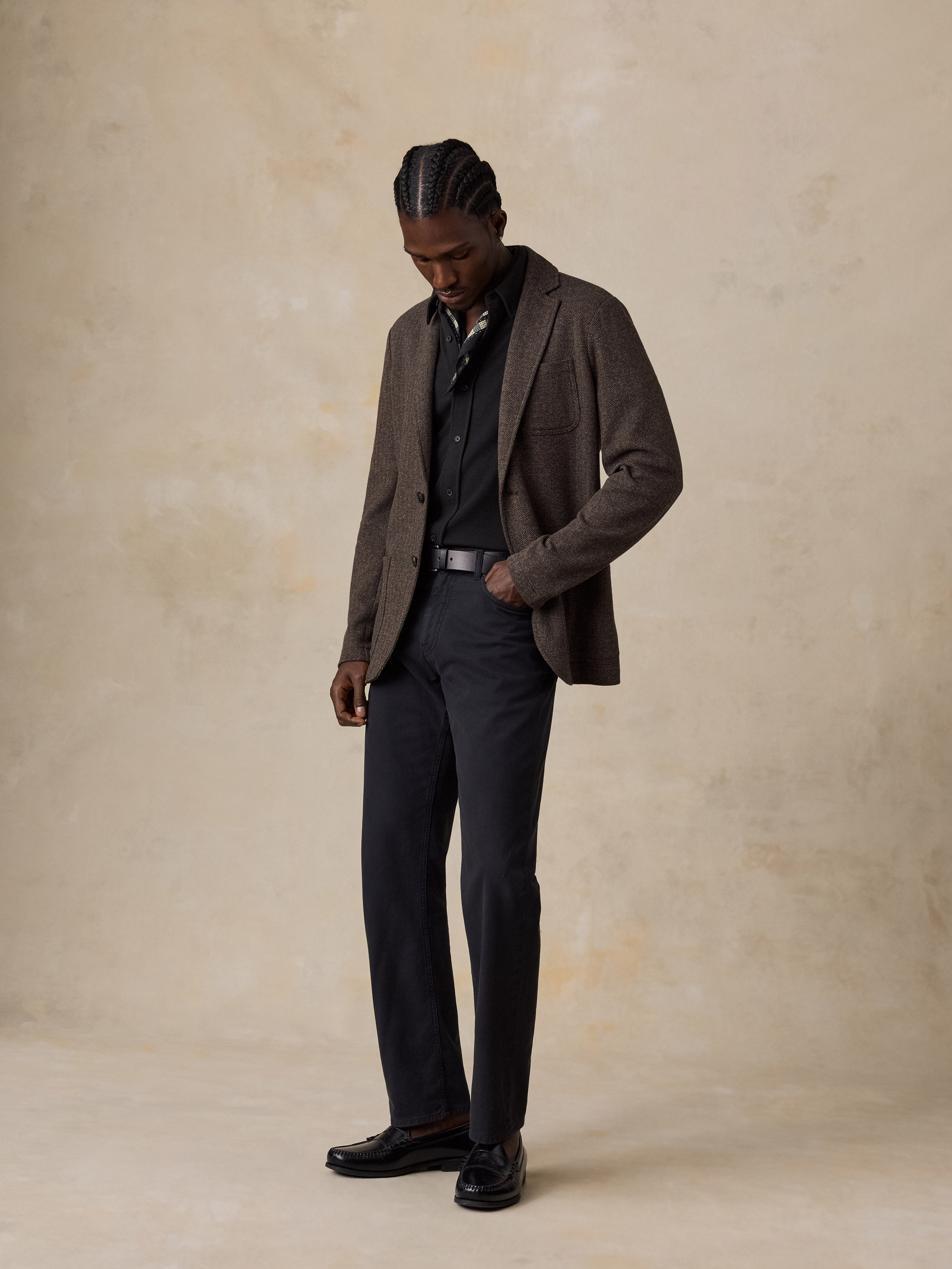 A man wearing a brown tweed blazer, dark navy trousers, a black shirt, and black loafers, standing against a textured beige studio backdrop.