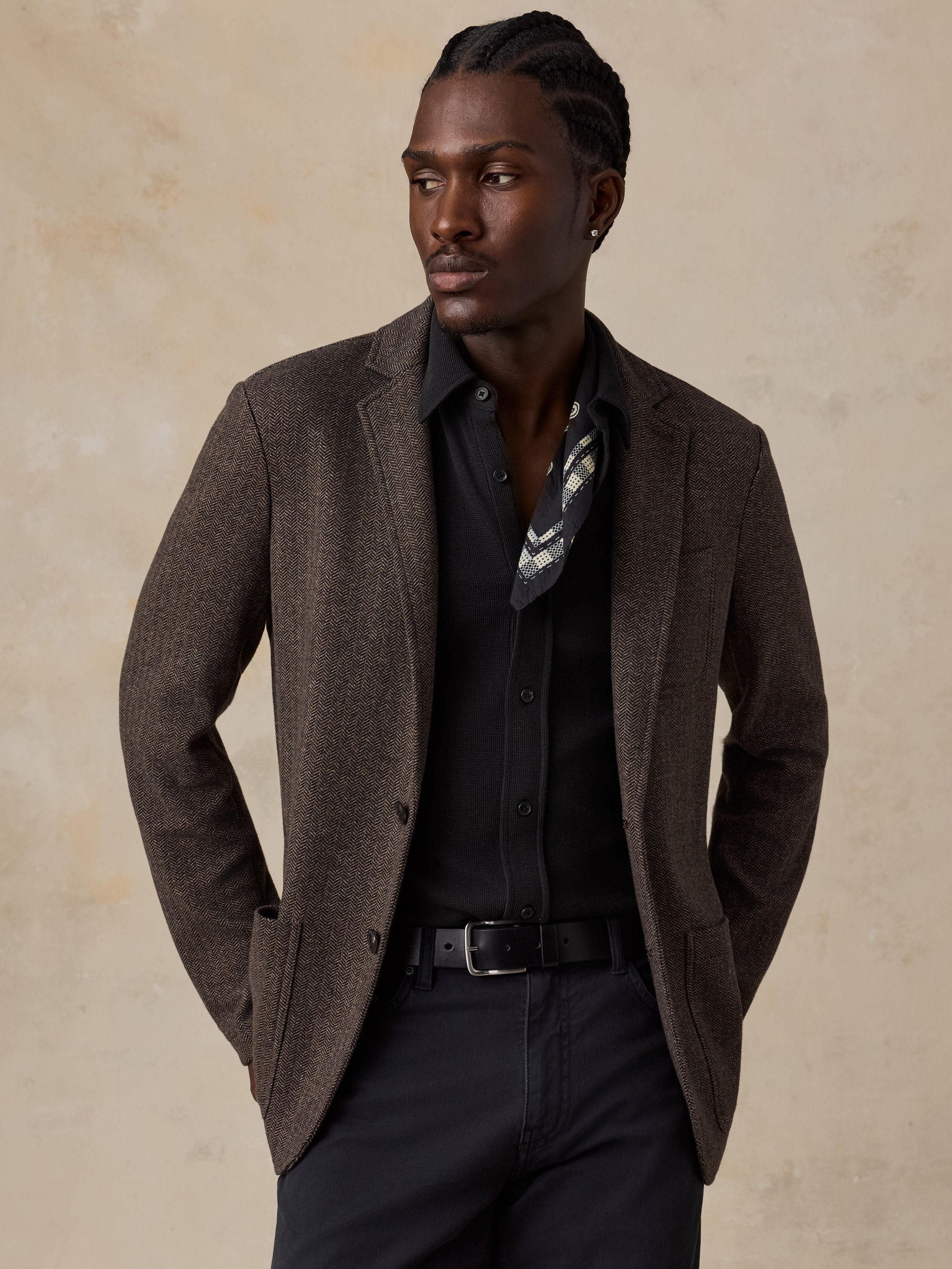 A close-up of a man wearing a brown herringbone blazer over a light blue button-down shirt, with his hands adjusting the blazer's front button.