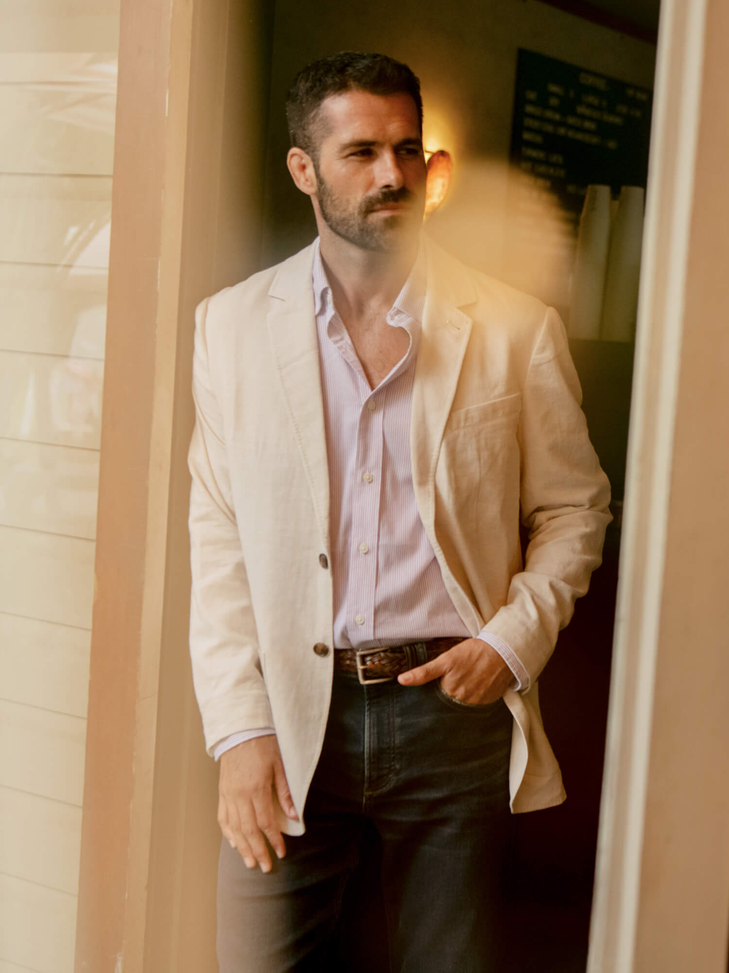 Man wearing a beige blazer over a purple shirt, standing indoors.