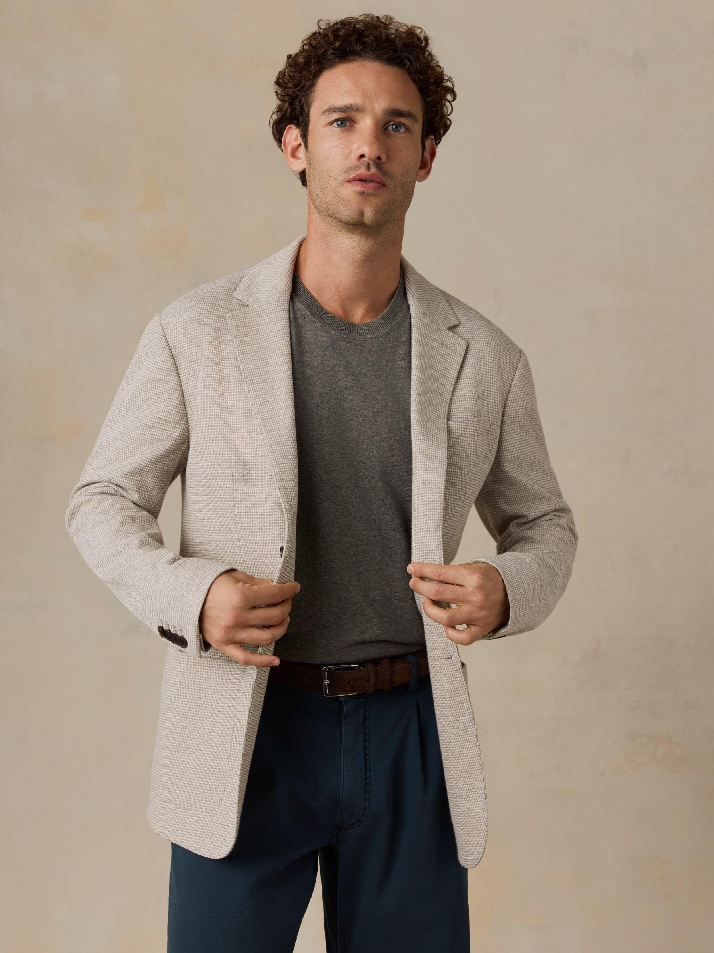 Man wearing a beige blazer over a gray shirt and dark pants on a beige background