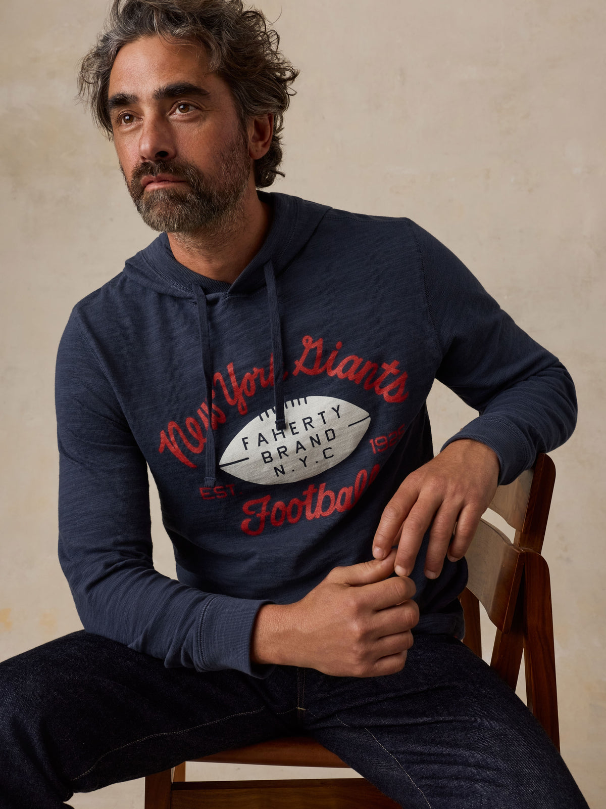 Man sitting on a chair wearing a navy NY Giants hoodie and blue jeans, image 6 of 6