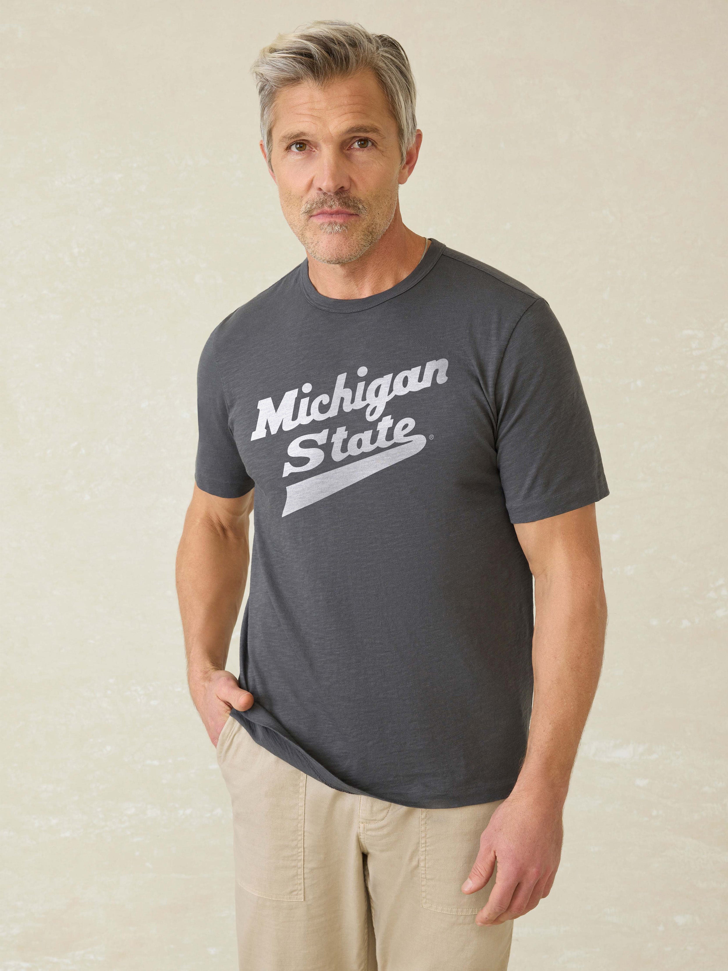 Man wearing a grey t-shirt with Michigan State graphic logo at the center front against a beige background