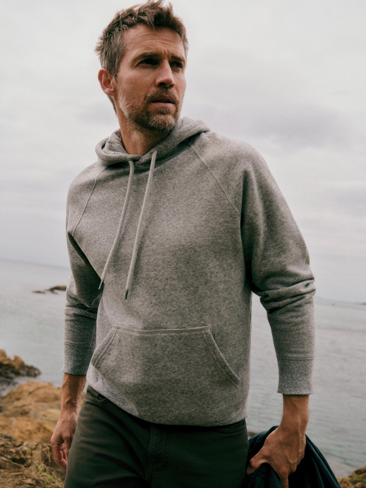 A man wearing a light gray heathered pullover hoodie with drawstrings and a kangaroo pocket, standing outdoors near the ocean under an overcast sky., image 1 of 7