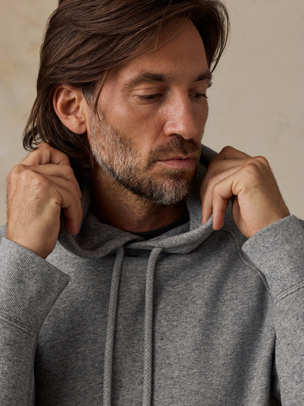 A close-up of a man wearing a heather gray pullover hoodie, adjusting the drawstrings around his neck, set against a neutral beige background., image 7 of 7