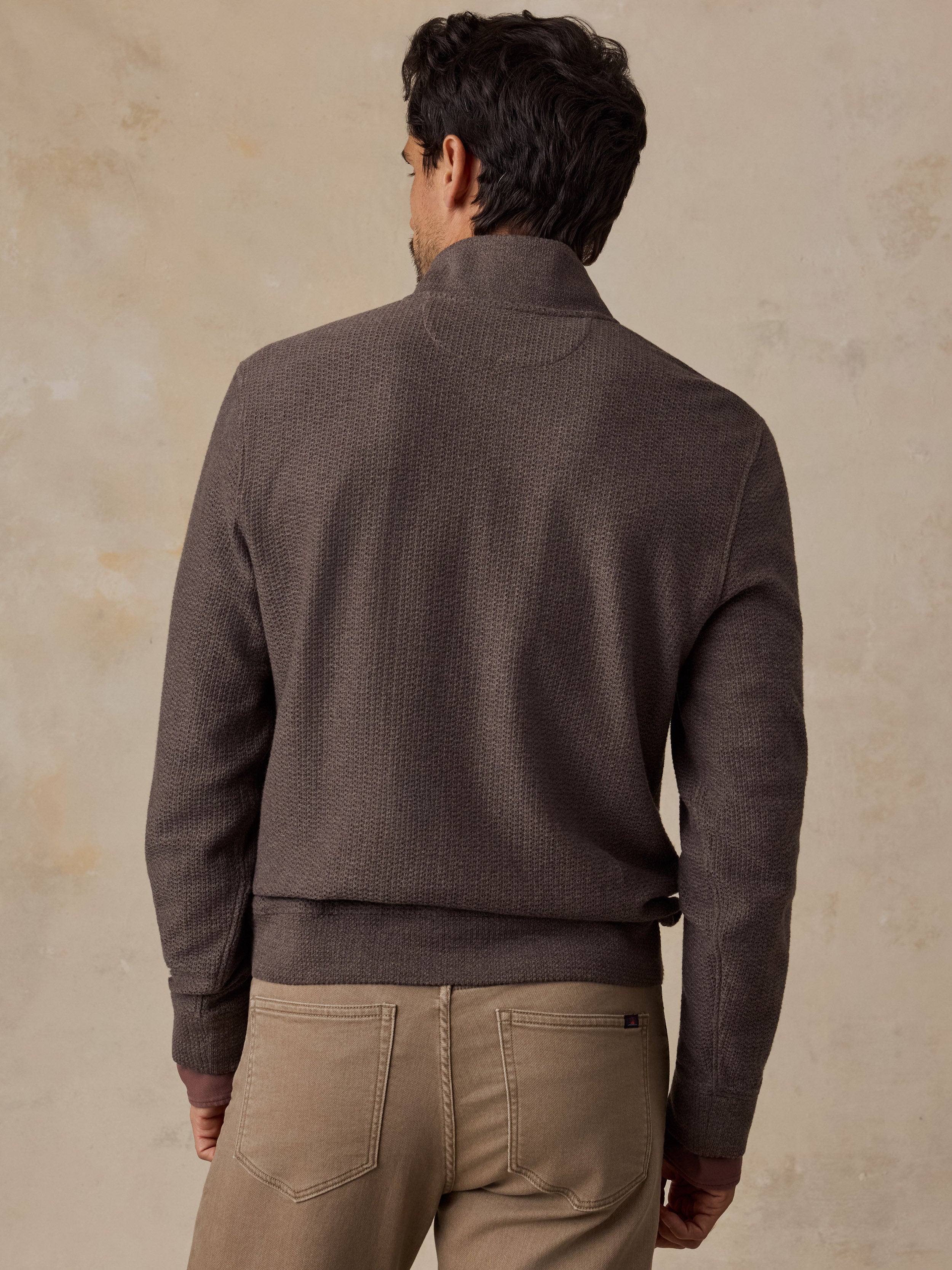 A man viewed from behind wearing a dark brown textured knit mock neck sweater and beige trousers, standing against a textured beige wall.