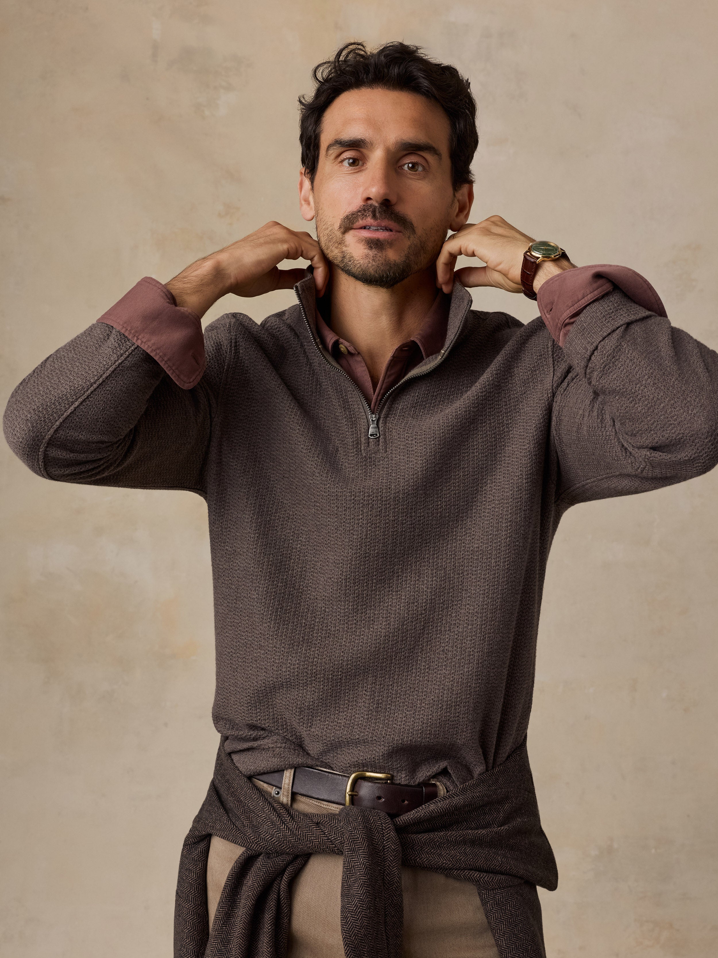 A man wearing a brown textured quarter-zip pullover sweater with contrasting dusty rose cuffs, styled with beige trousers and a brown belt, posing against a neutral textured beige background.