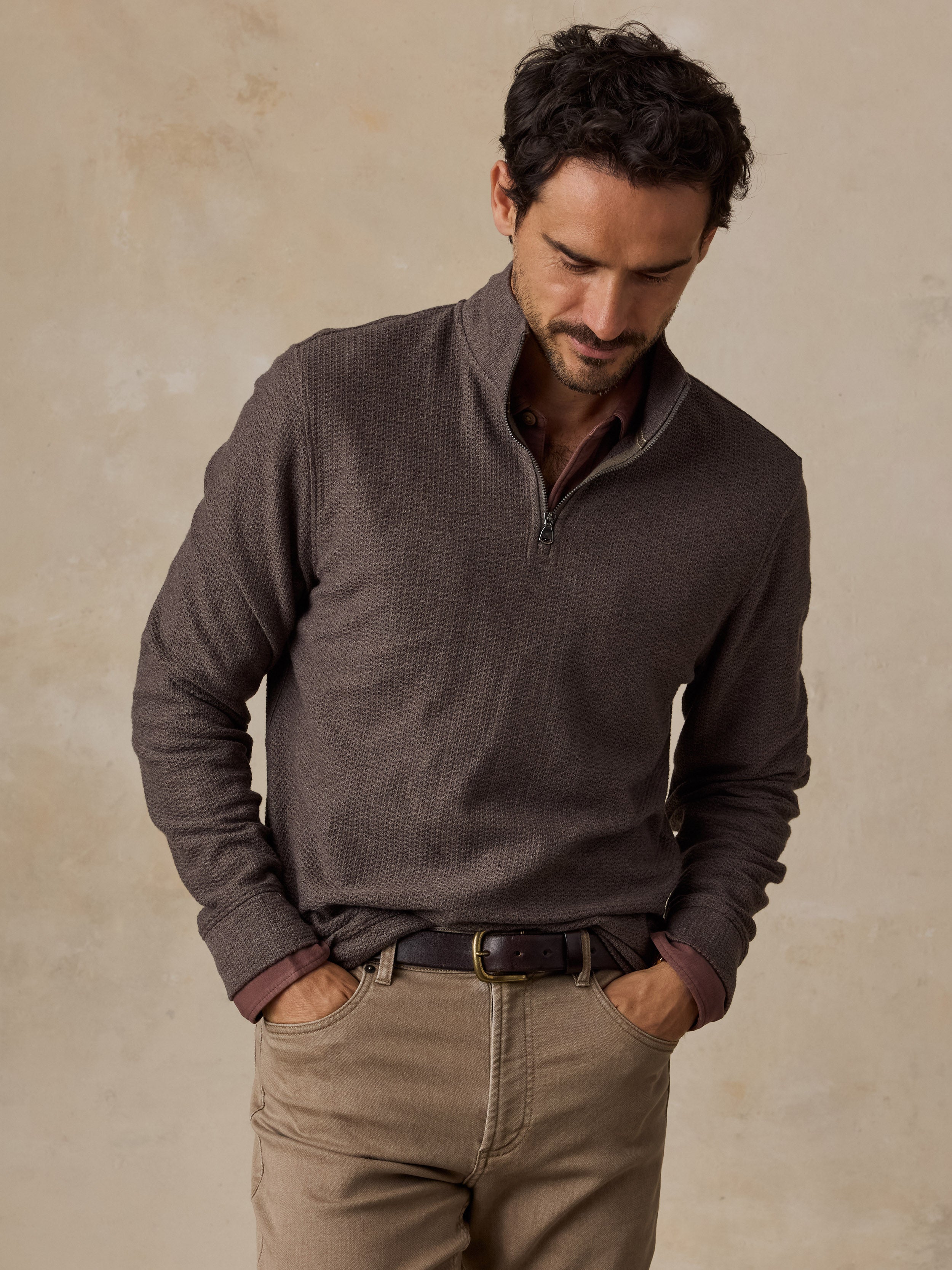 A man wearing a dark brown waffle-knit quarter-zip pullover sweater and khaki pants, standing against a textured beige background.