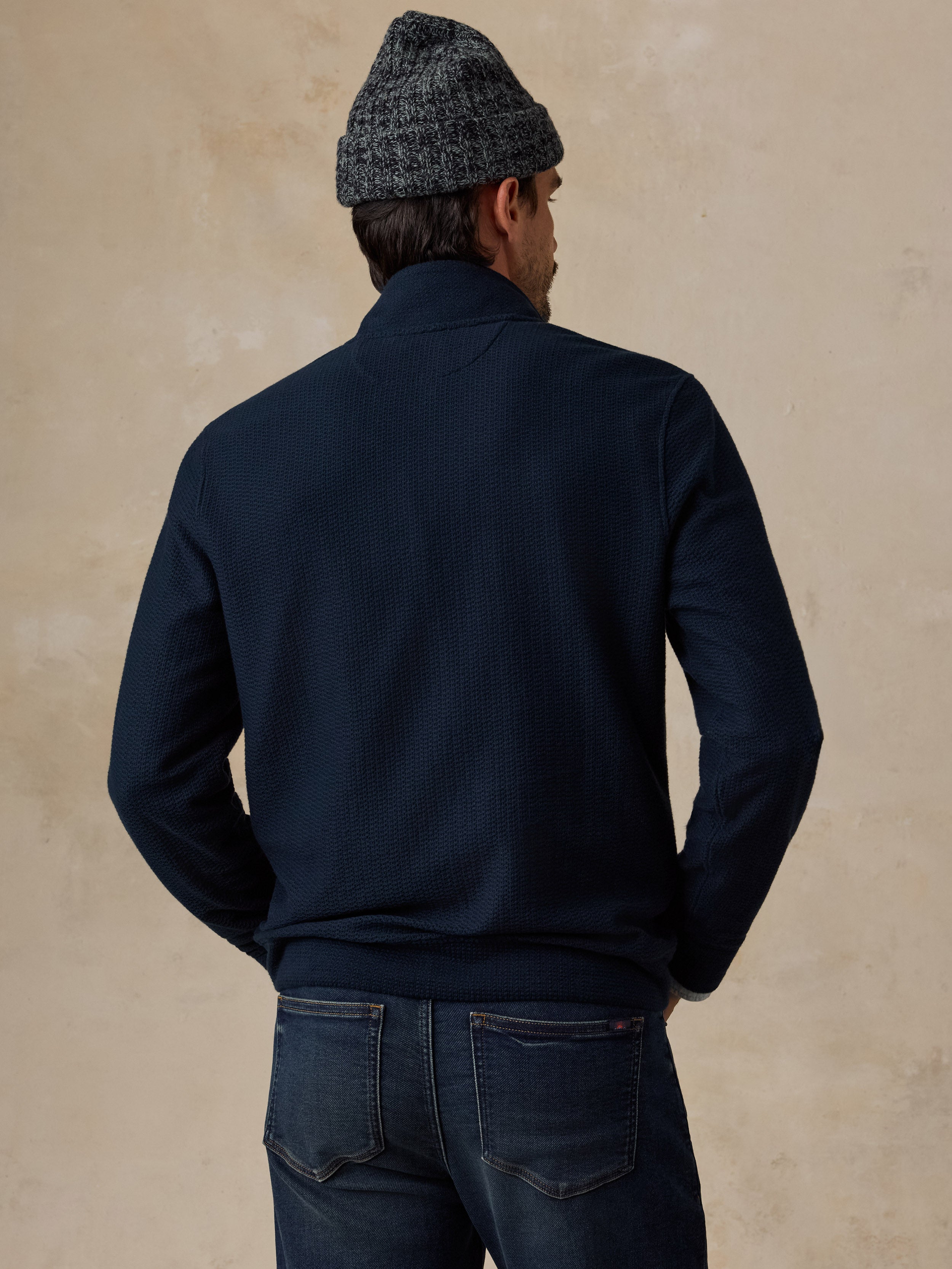 A man viewed from behind wearing a dark navy blue knit sweater, a gray and black patterned beanie, and dark blue jeans, standing against a textured beige wall.