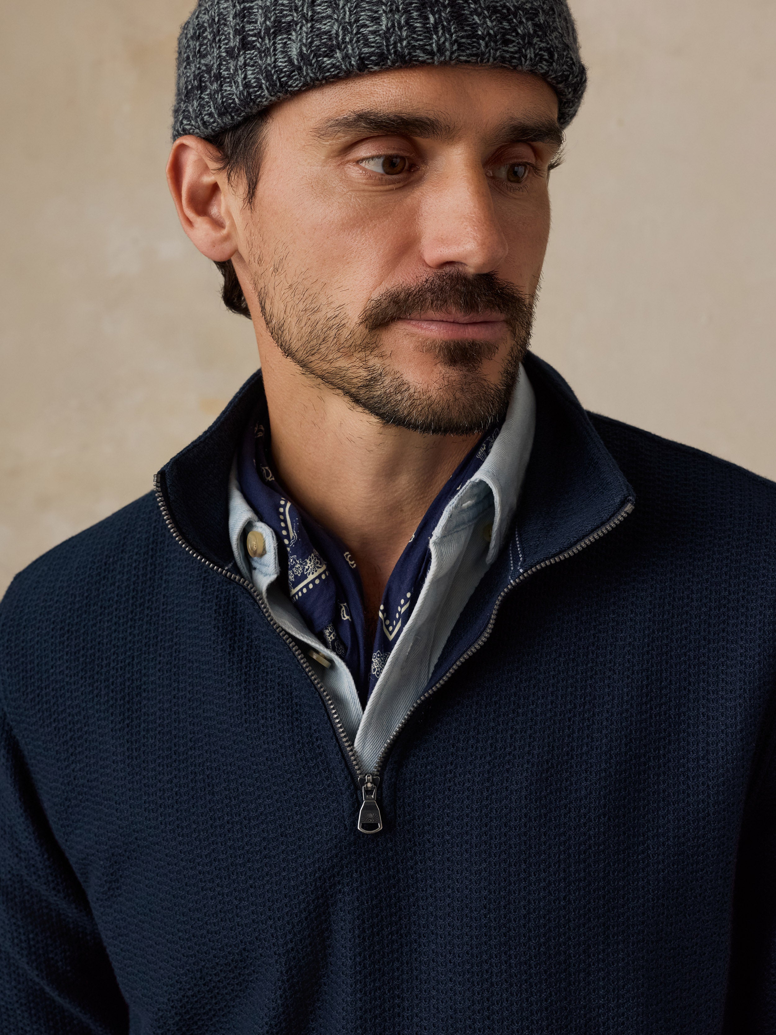 A close-up of a man wearing a dark navy quarter-zip sweater over a light blue collared shirt with a patterned neck gaiter, paired with a gray knit beanie, set against a warm beige background.