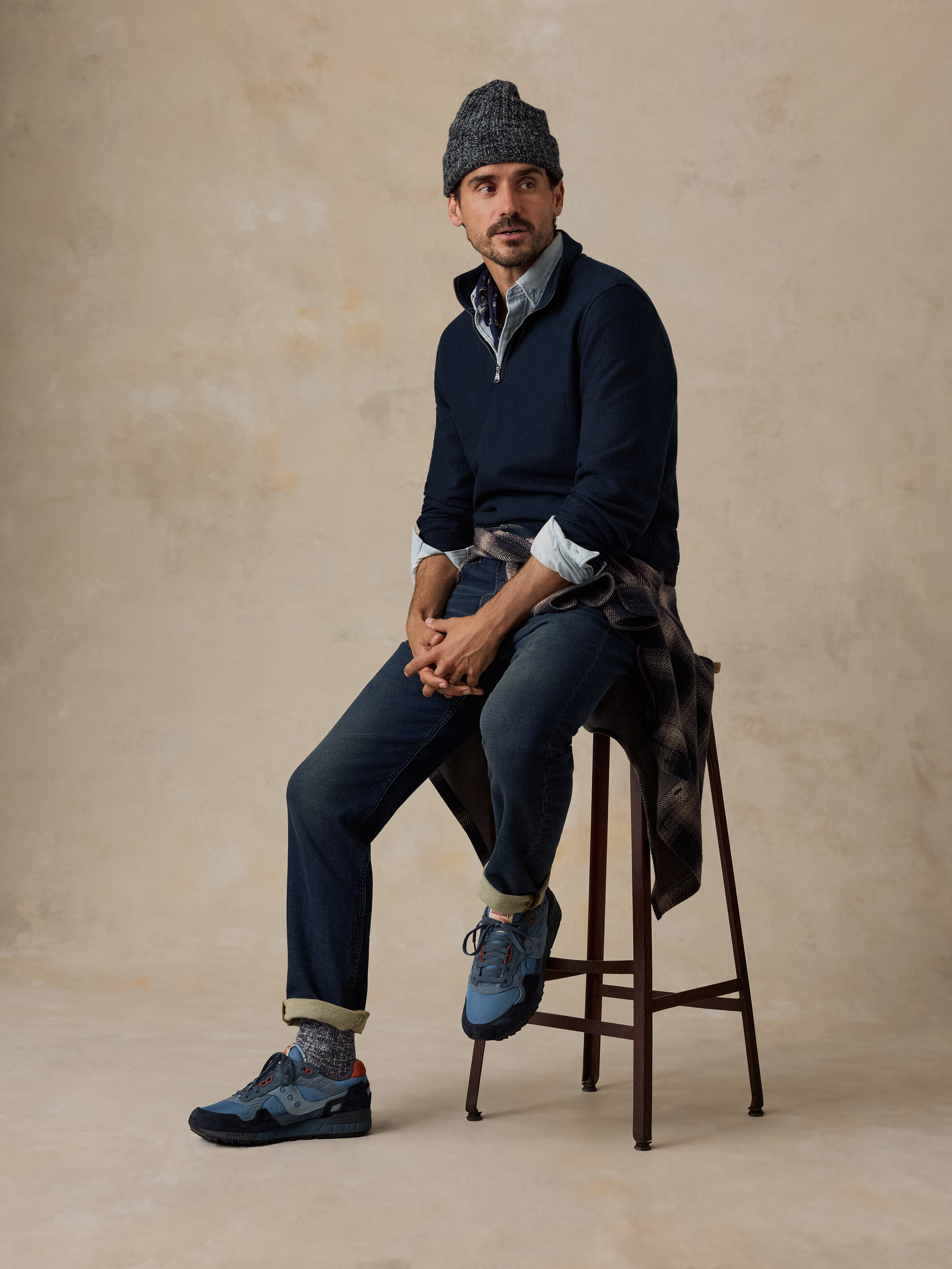 A man sits on a wooden stool wearing a navy quarter-zip sweater, blue jeans, blue sneakers, a patterned knit beanie, and a plaid shirt tied around his waist, set against a textured beige studio backdrop.