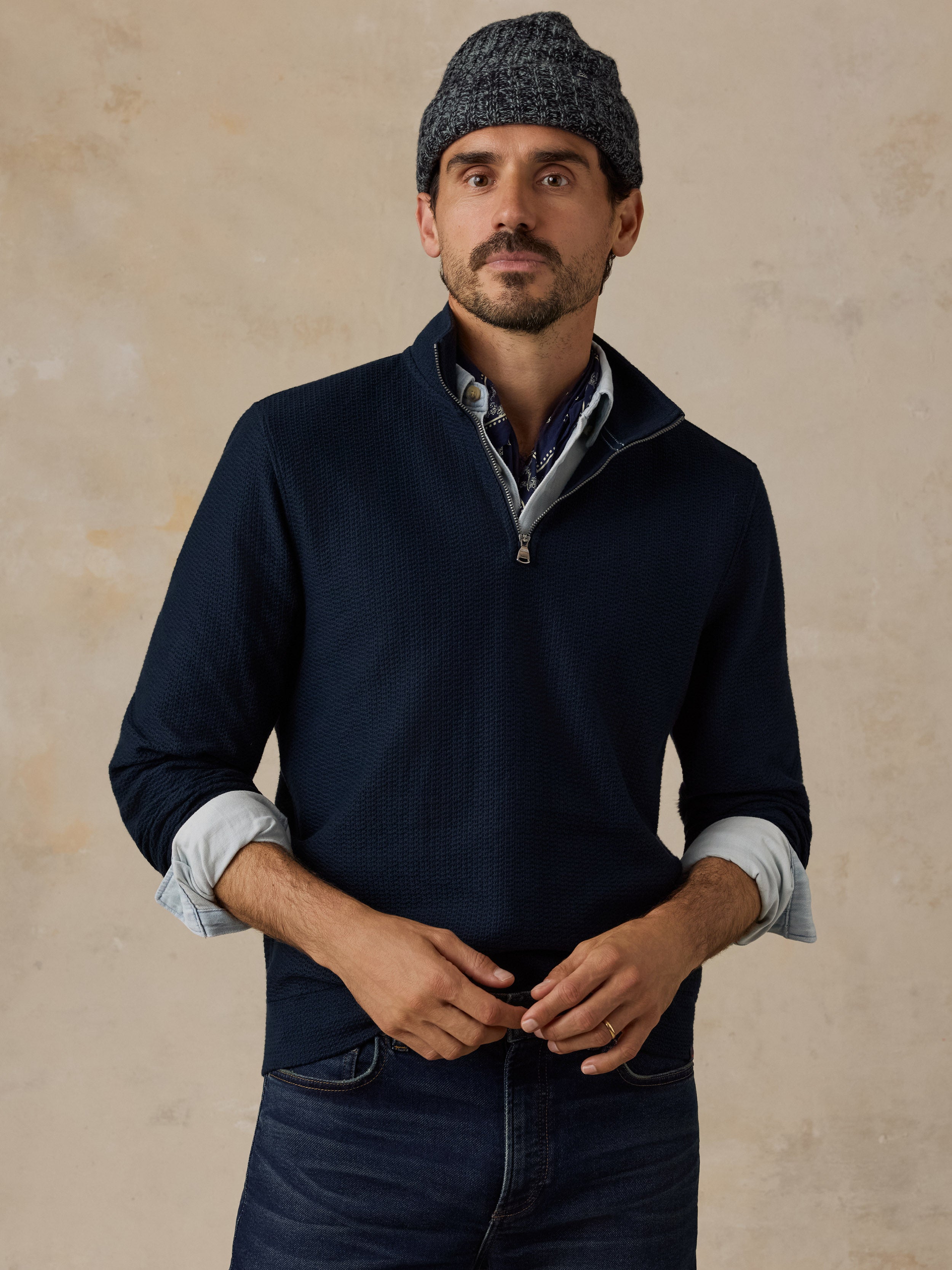 A man wearing a navy blue quarter-zip sweater over a light blue collared shirt, paired with dark blue jeans and a dark gray knit beanie, standing against a textured beige background.