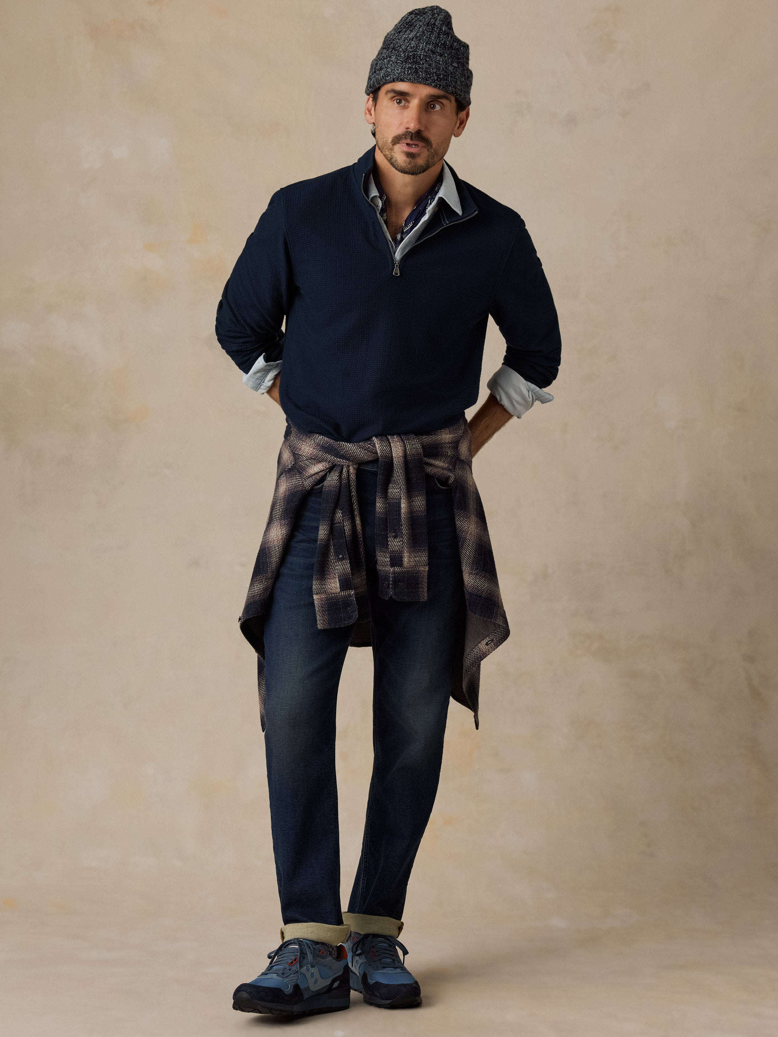 A man modeling a dark navy quarter-zip sweater vest over a light blue collared shirt, paired with dark blue jeans, a gray knit beanie, and a brown plaid shirt tied around his waist, set against a textured beige studio backdrop.