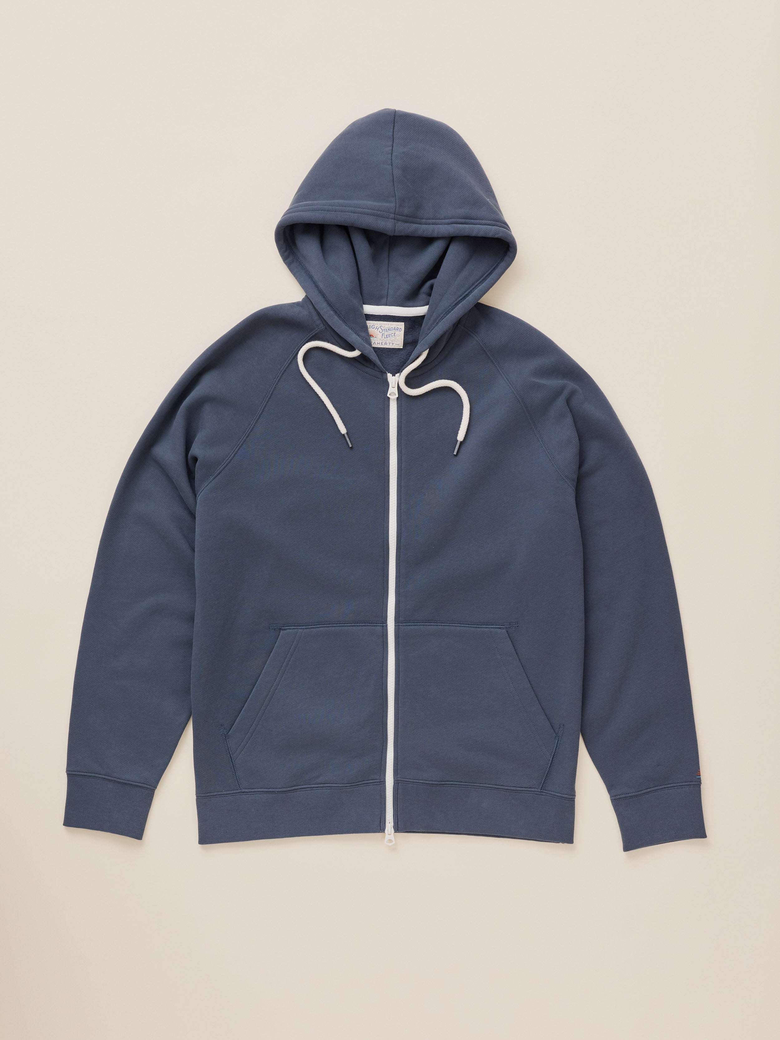 A slate blue zip-up hoodie with white drawstrings and a white front zipper, laid flat against a light beige background.