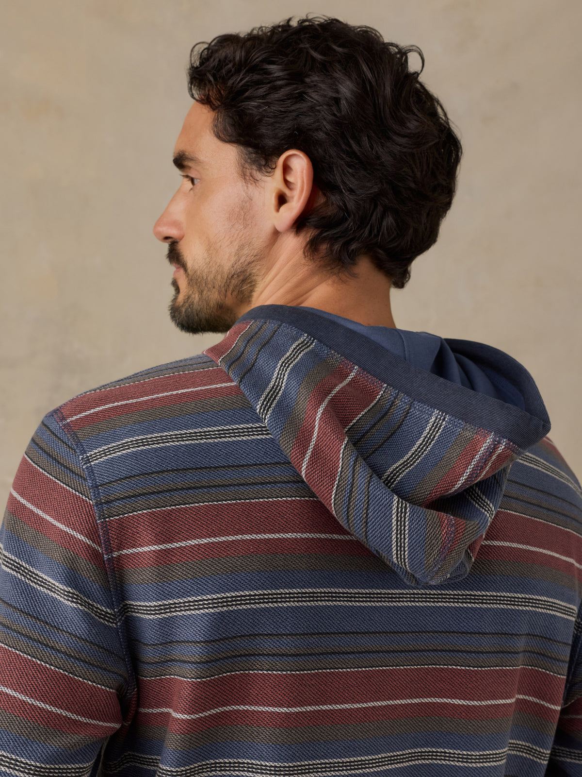 A man shown from the back wearing a striped pullover hoodie with horizontal bands of navy blue, rust red, and cream., image 6 of 7
