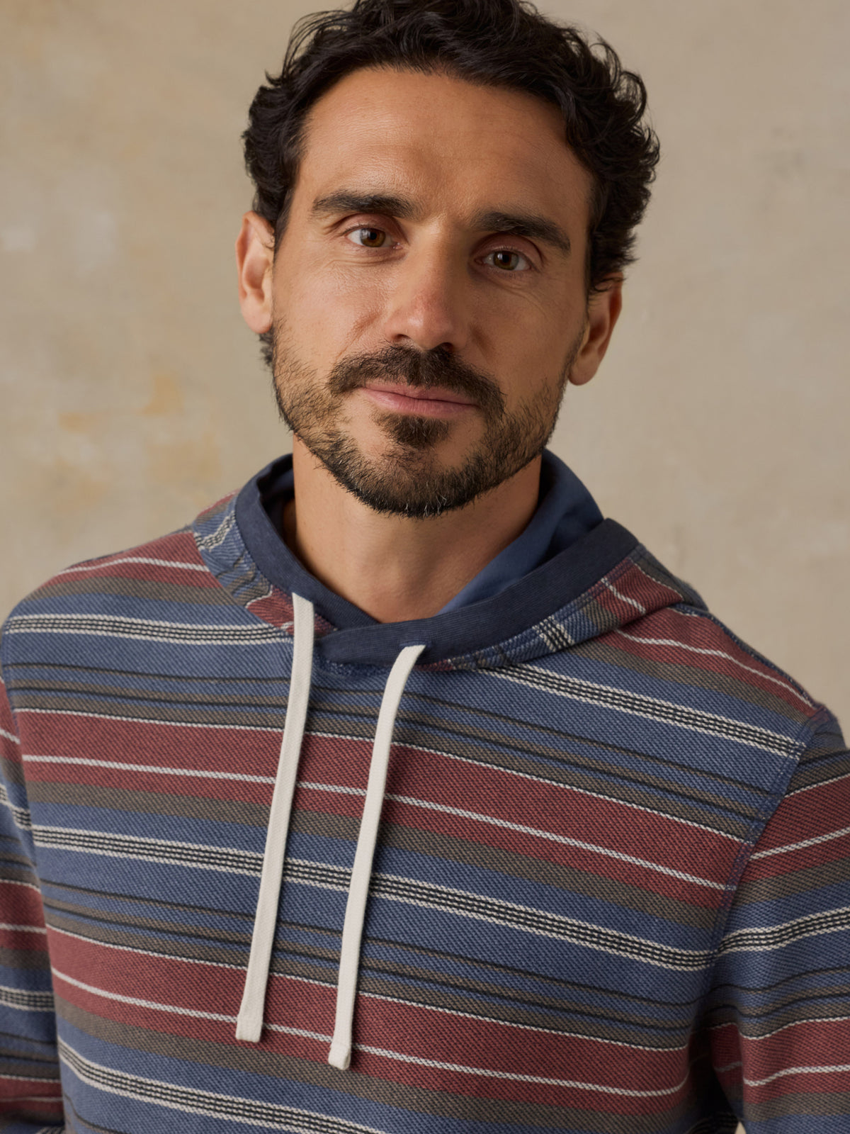 A man wearing a blue, red, and beige striped hoodie with white drawstrings, photographed against a textured beige background., image 4 of 7