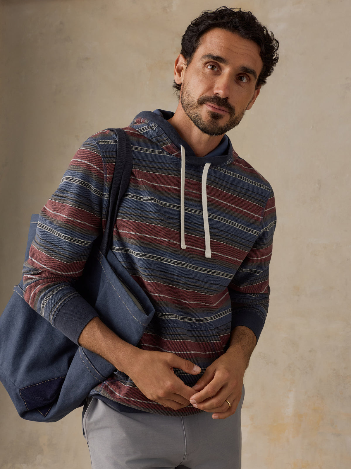 A man wearing a blue, red, and beige striped hoodie with white drawstrings, carrying a navy blue tote bag over his shoulder, set against a textured beige wall., image 7 of 7