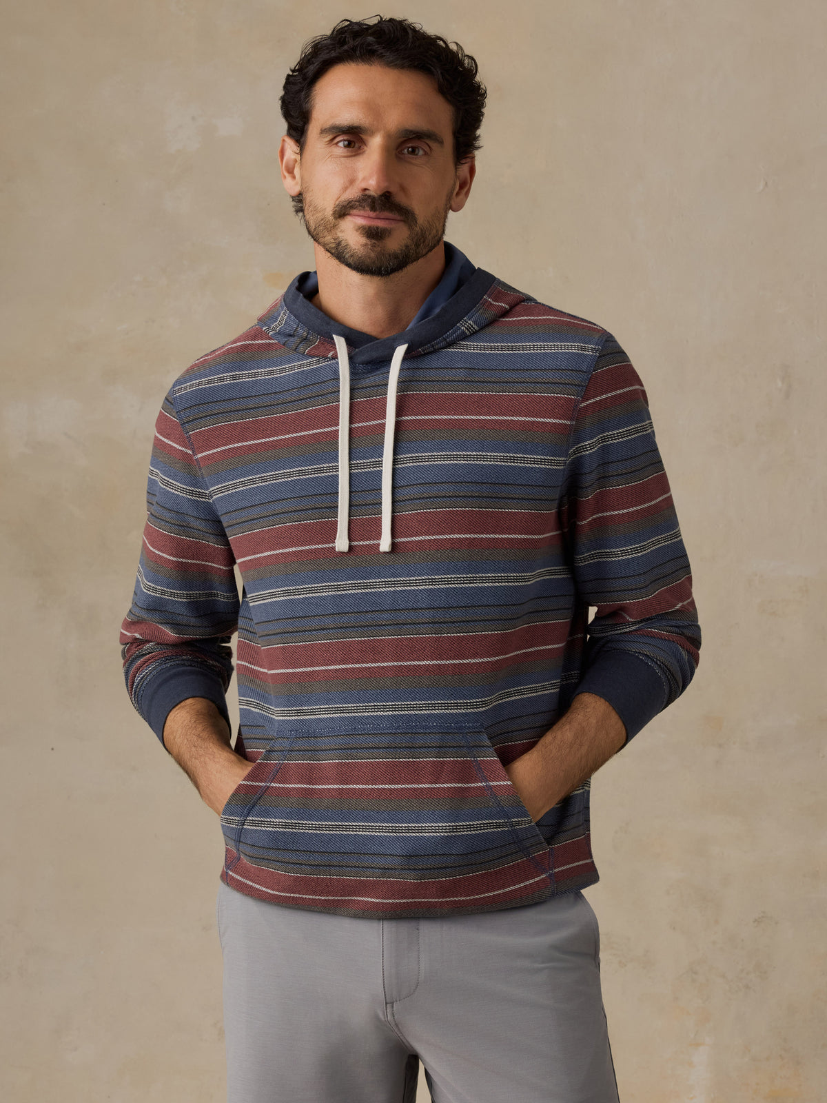 A man wearing a long-sleeve striped hoodie with blue, red, and beige horizontal stripes, paired with light gray trousers, standing against a textured beige wall., image 2 of 7