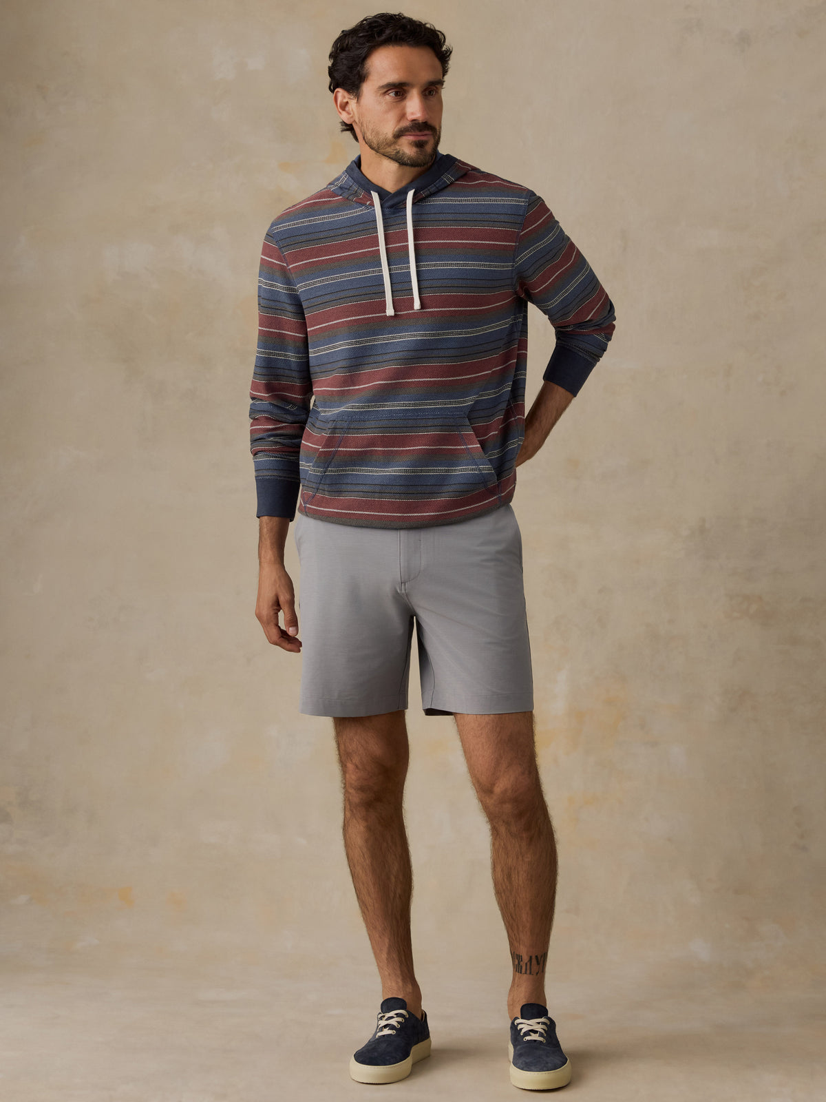 A man wearing a long-sleeve striped hoodie with navy, red, and gray horizontal stripes, white drawstrings, gray shorts, and navy blue sneakers, standing against a textured beige studio backdrop., image 5 of 7