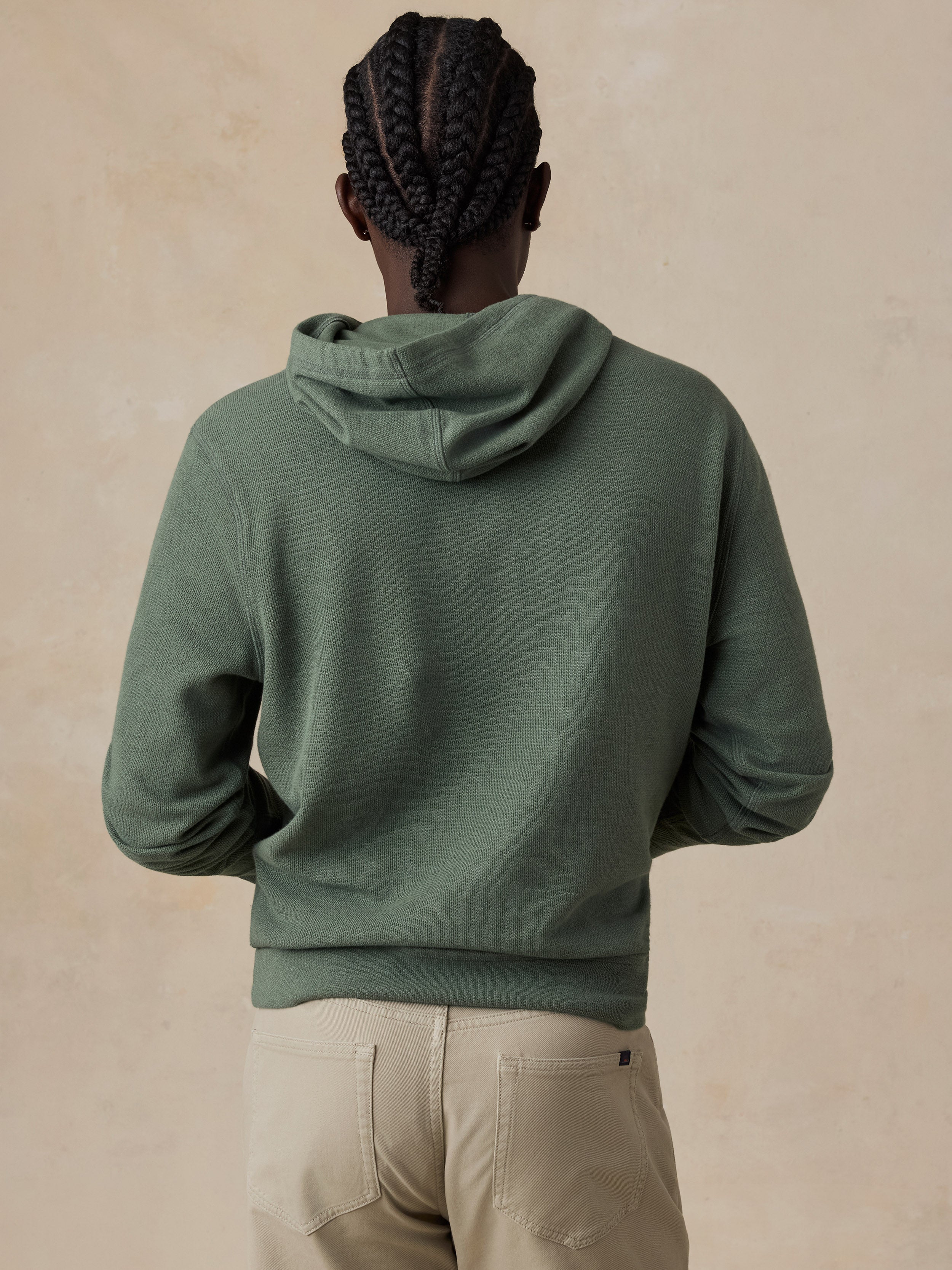 A man shown from the back wearing an olive green hooded sweatshirt and beige pants, standing against a textured beige wall.