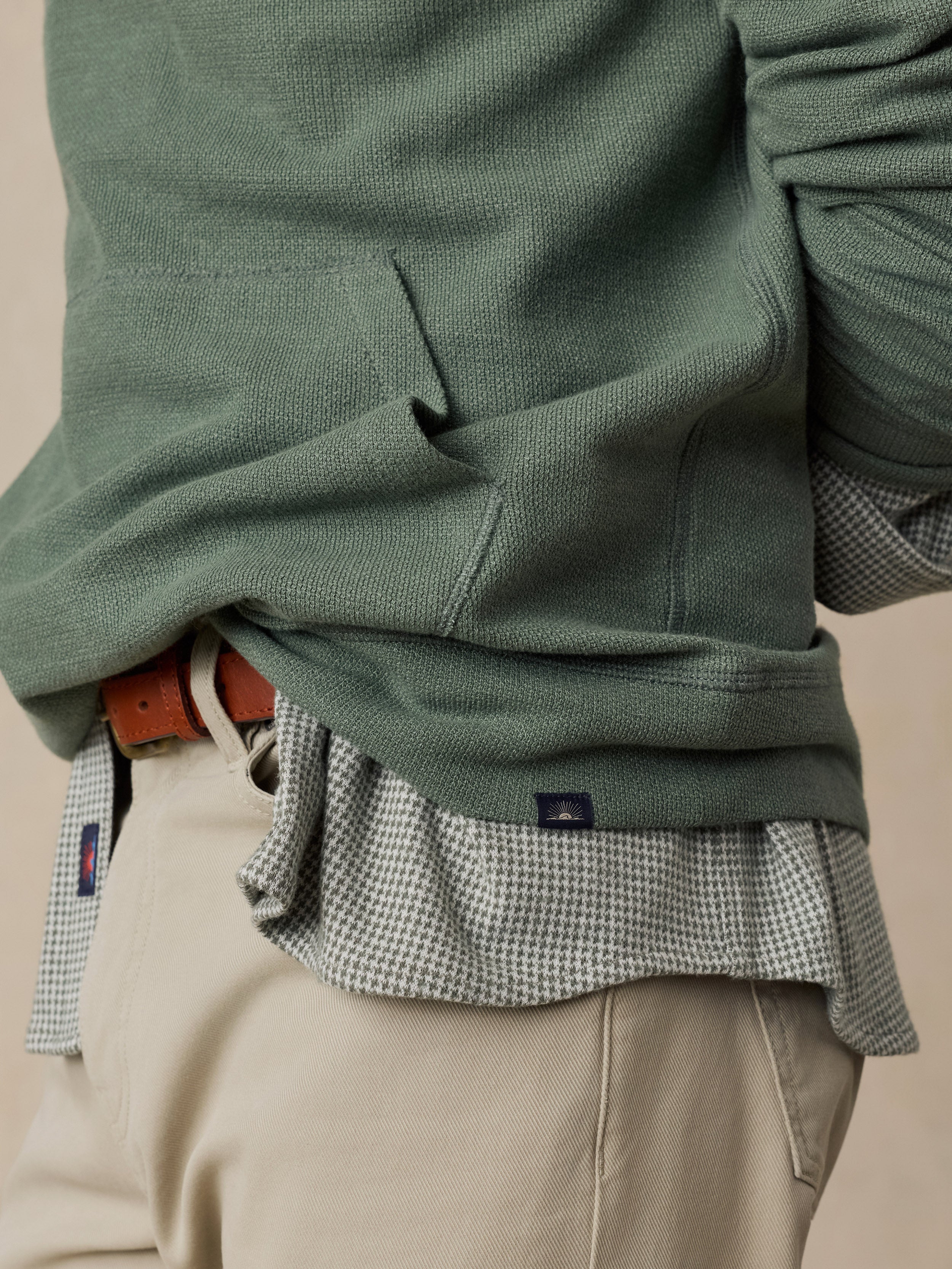 Close-up of a man wearing a sage green knit sweater layered over a small-scale checkered shirt and beige trousers, with a brown belt visible.