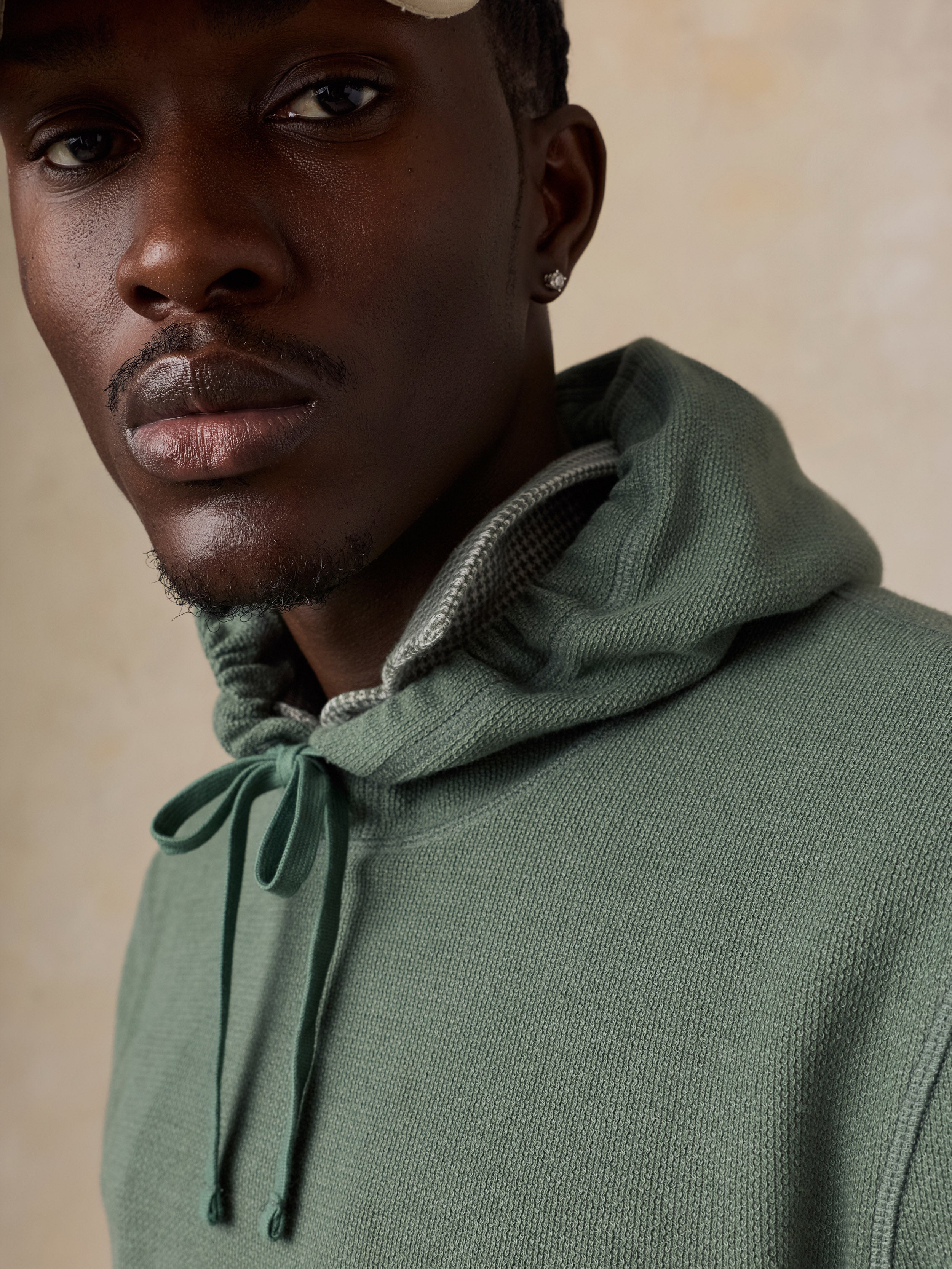 A close-up shot of a man wearing a sage green knit hoodie with a gray patterned inner hood lining and matching drawstrings, set against a warm beige background.