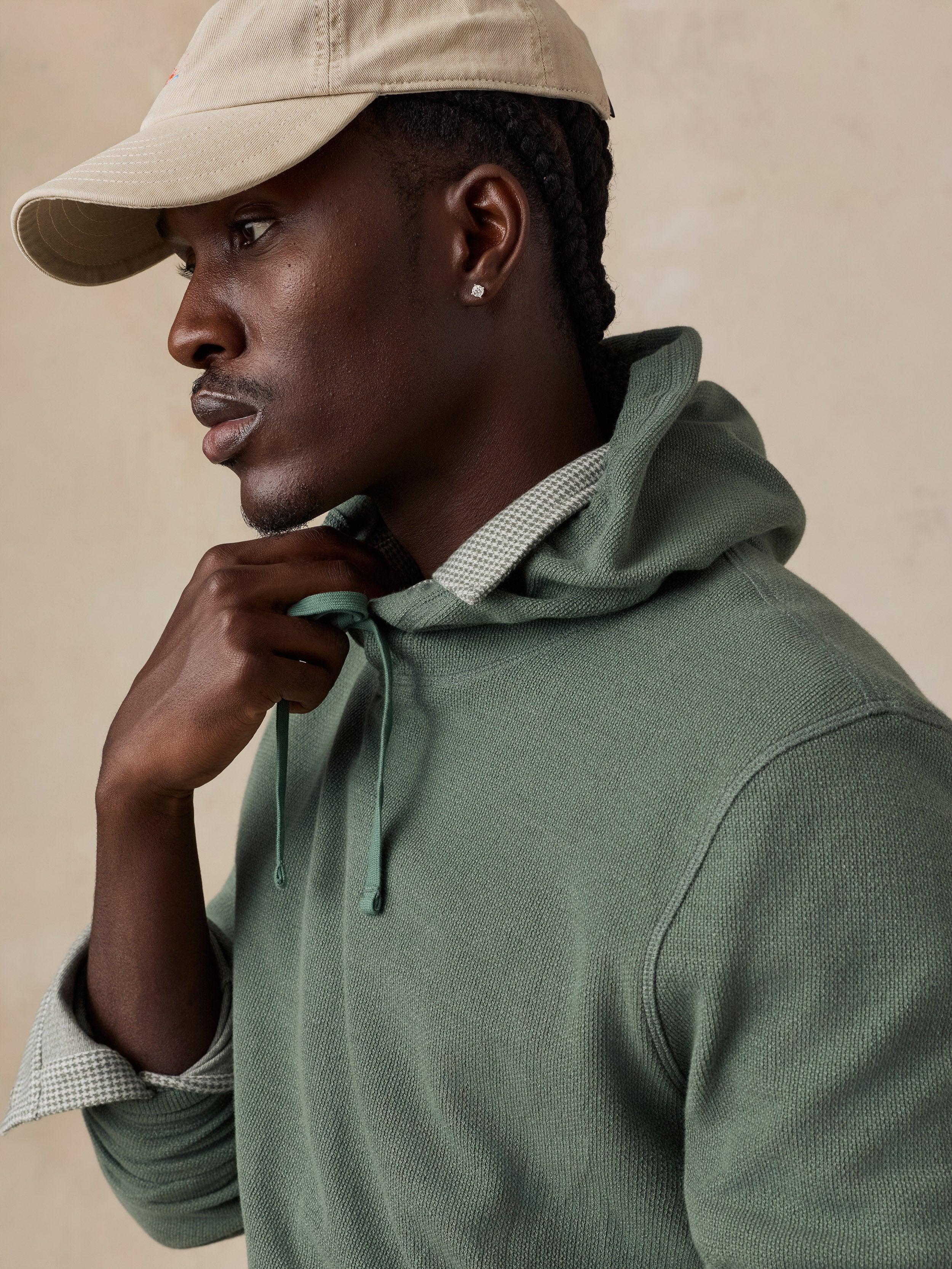 A man wearing an olive green knit hoodie with a matching drawstring, layered over a light-colored checkered shirt, and wearing a beige baseball cap, set against a warm beige background.