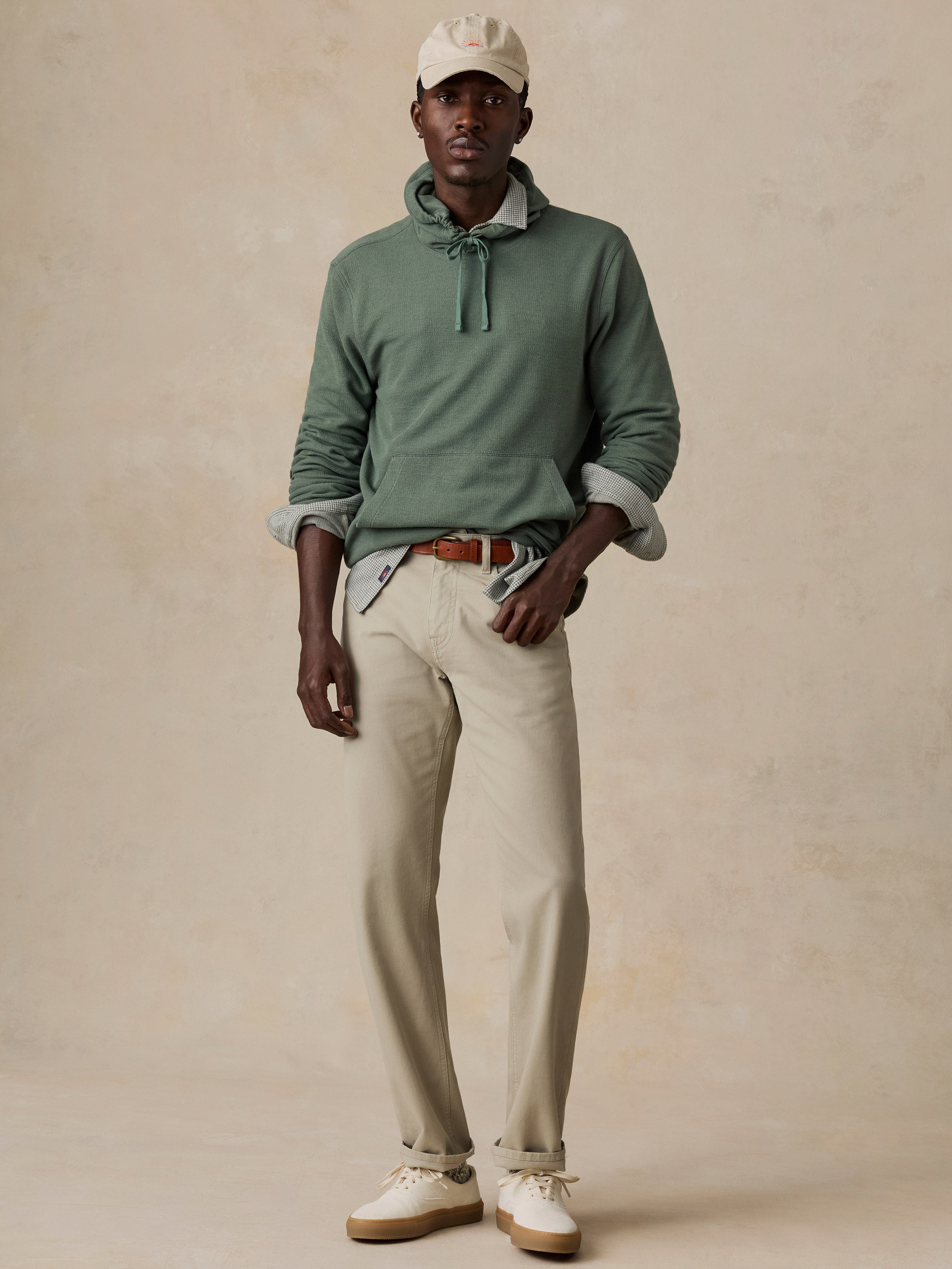 A man modeling a sage green knit pullover sweater with a tie-neck detail, styled with beige trousers, a brown leather belt, a light green shirt underneath, a cream cap, and white sneakers against a textured beige studio backdrop.